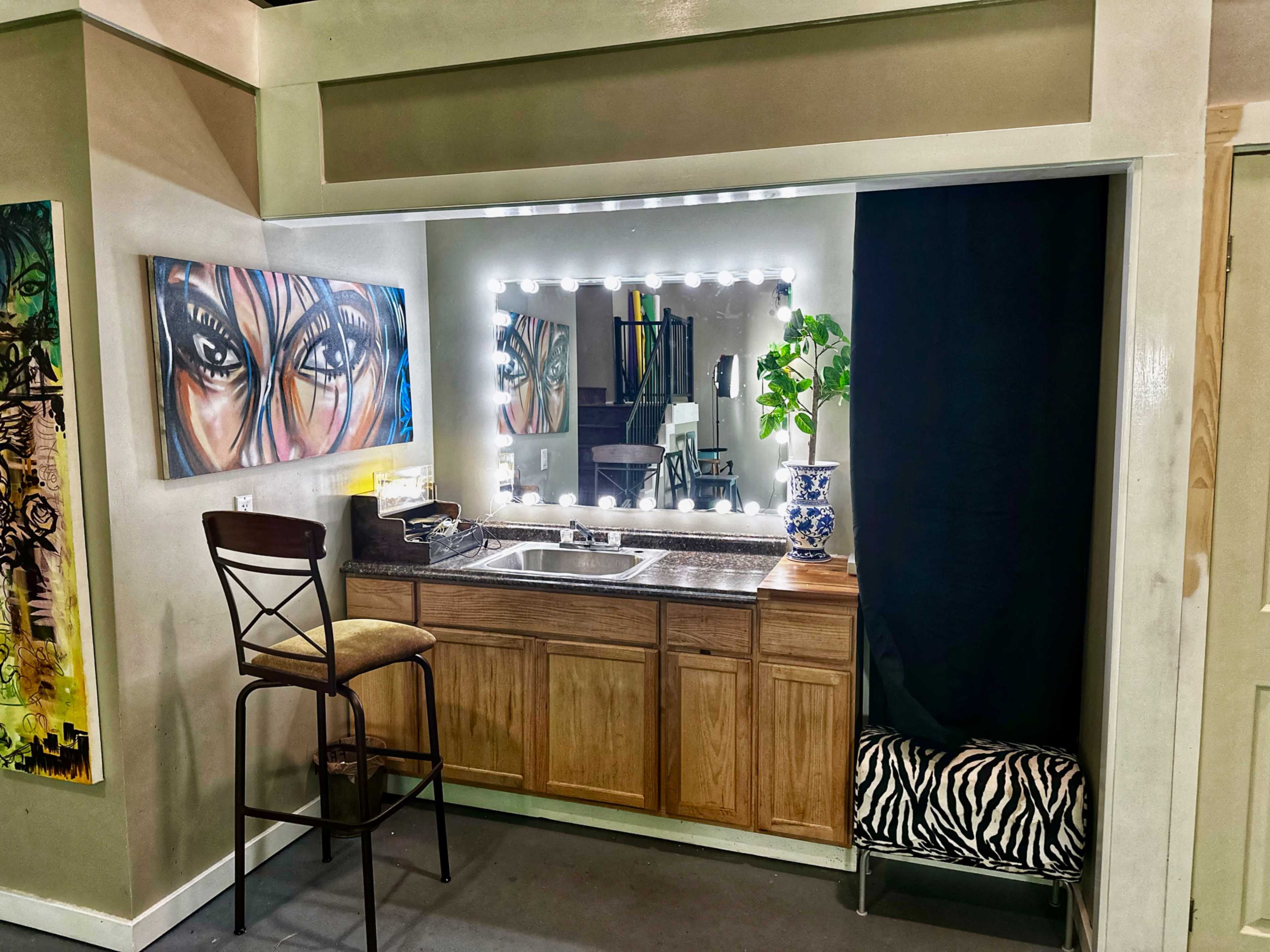The image shows a well-lit vanity area with a mirror, a wooden countertop, a chair, and a decorative plant, adjacent to an artistic painting on the wall.