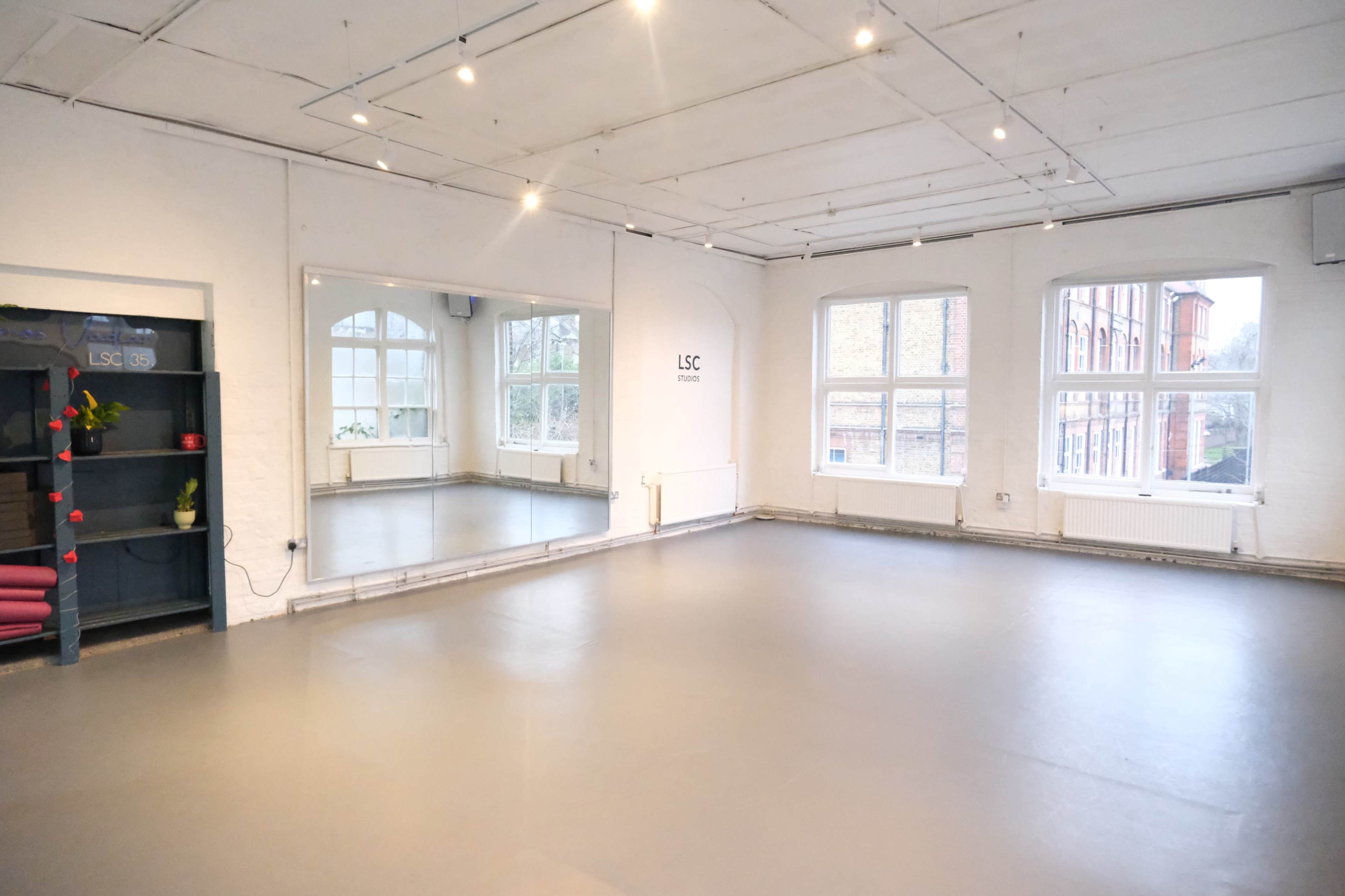The image shows an empty studio space with a large mirror on one wall, large windows allowing natural light, and a shelf with colorful items in the corner.