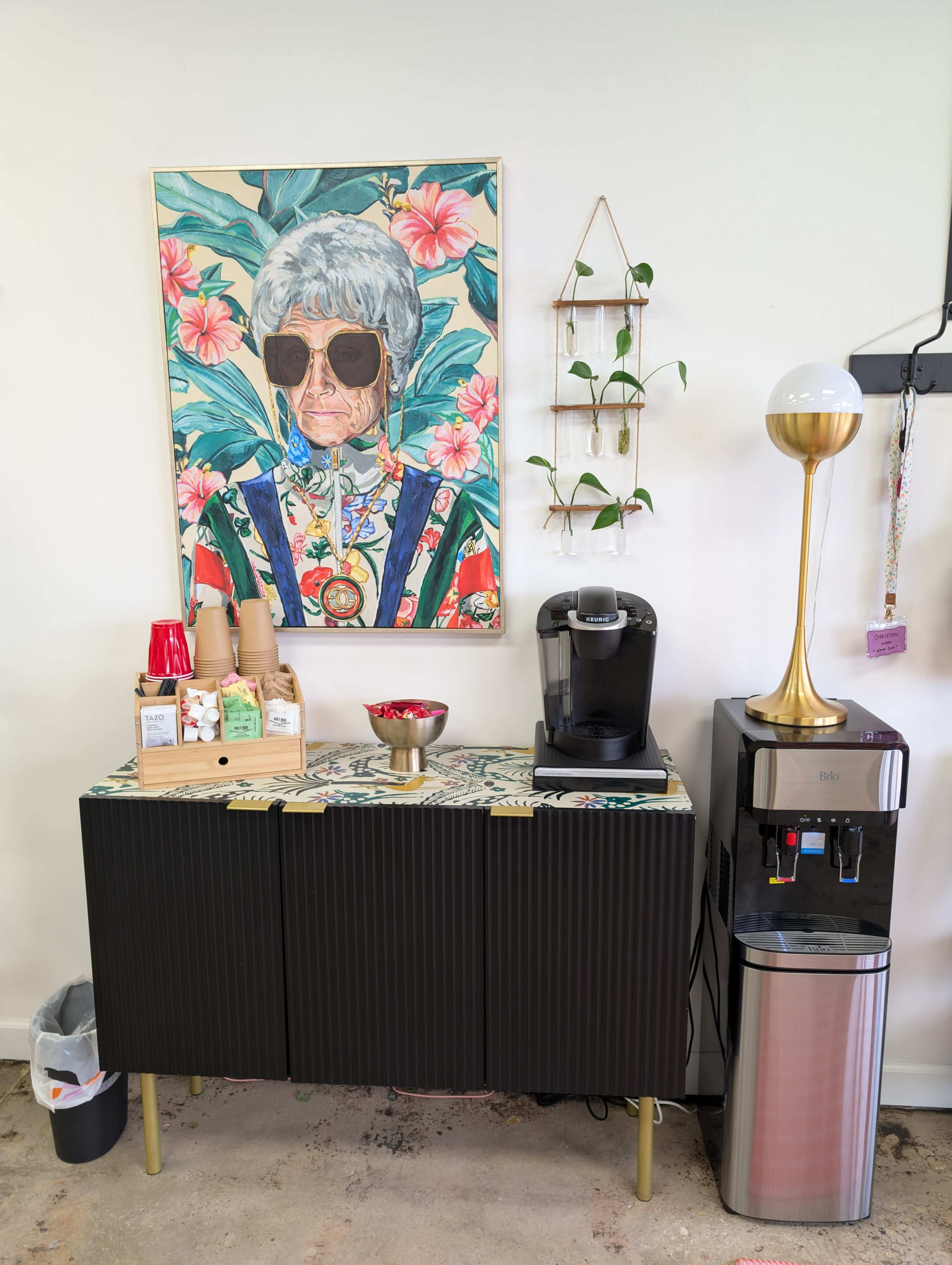 A coffee station featuring a portrait of a woman in sunglasses, a coffee maker, and a water dispenser against a wall adorned with plants.