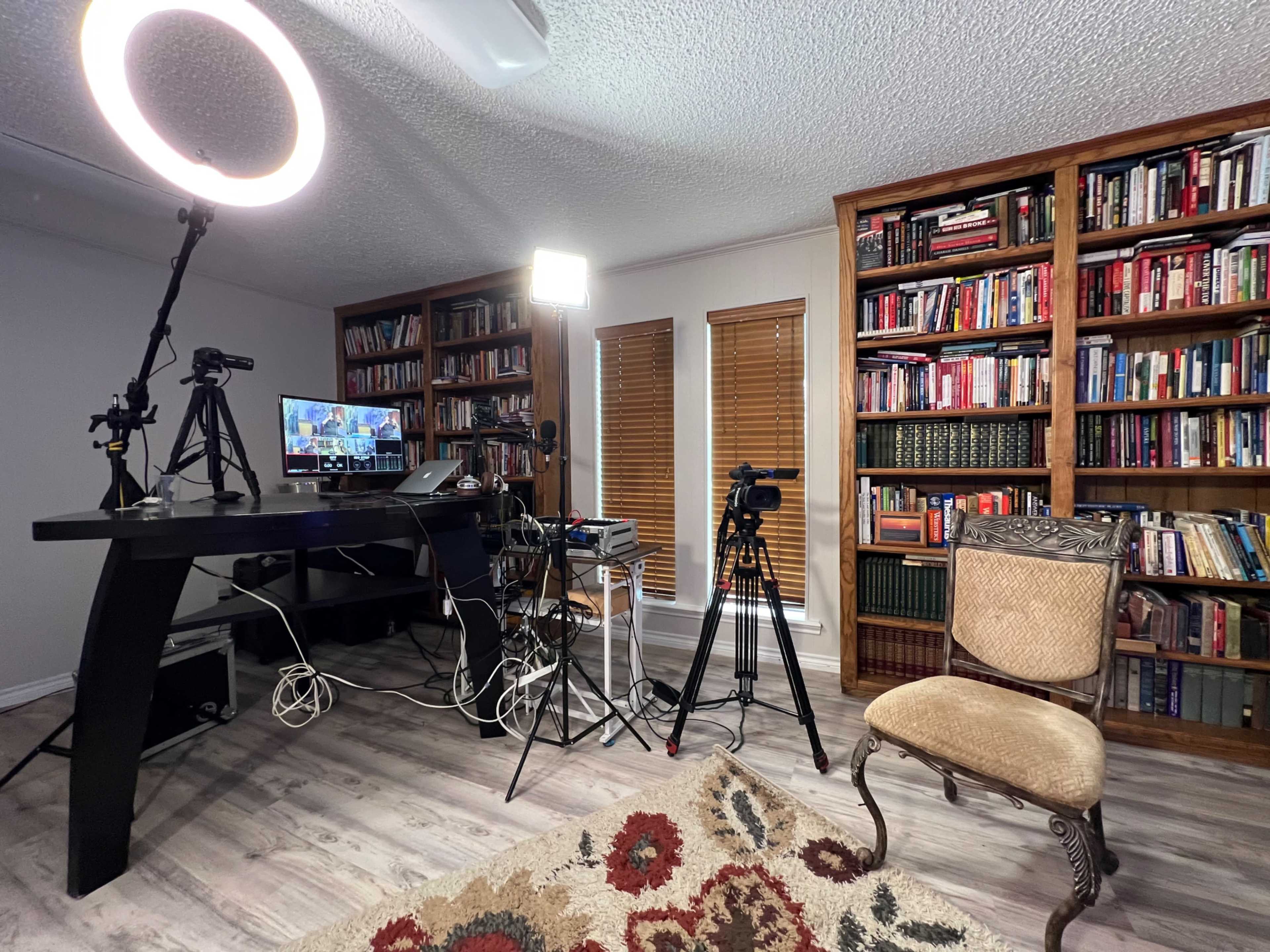 A room with a desk, multiple cameras, and light equipment set up next to a bookshelf filled with books, with an empty chair positioned nearby.