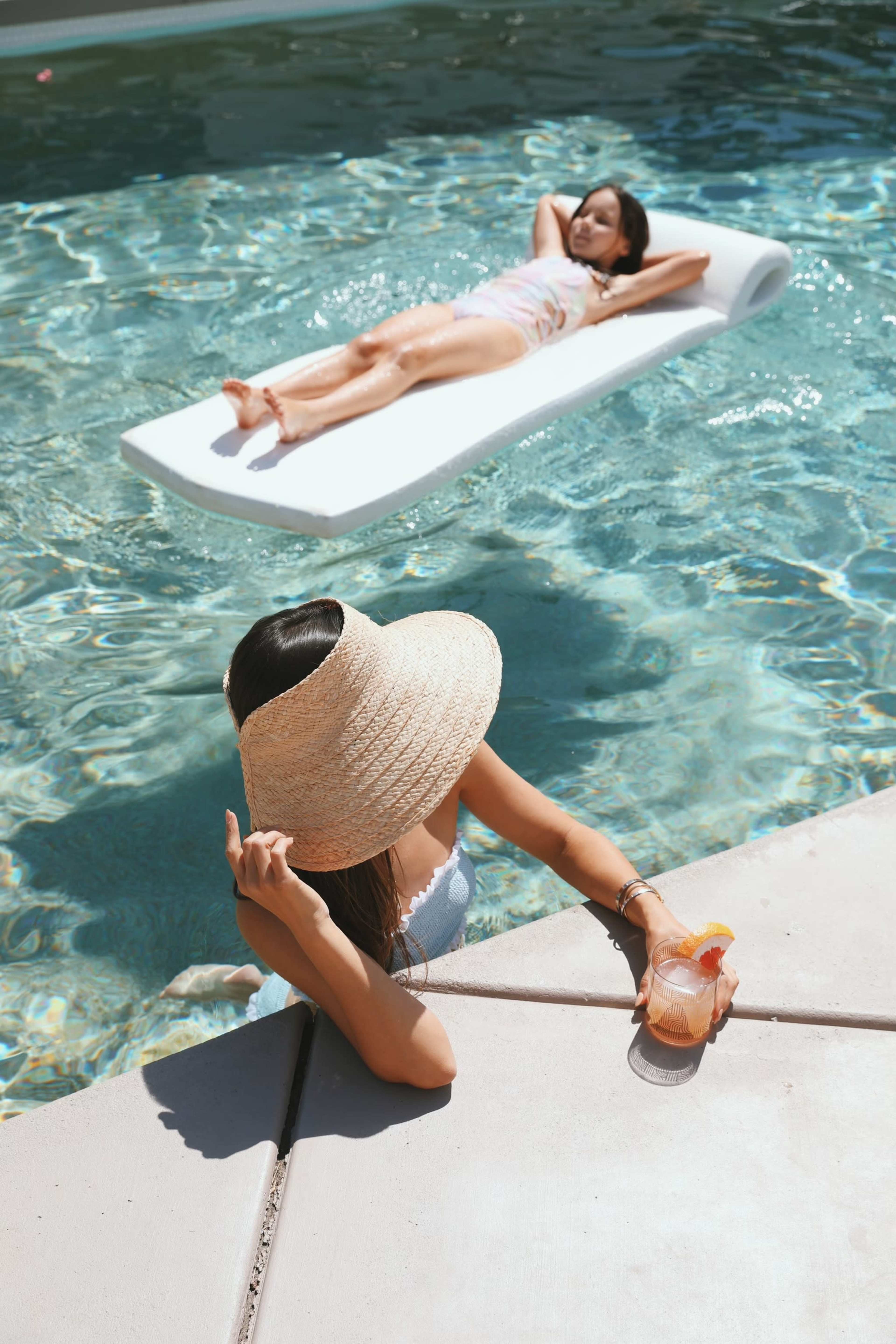 Two women relax by a pool, one floats on a raft while the other sits at the edge with a drink.