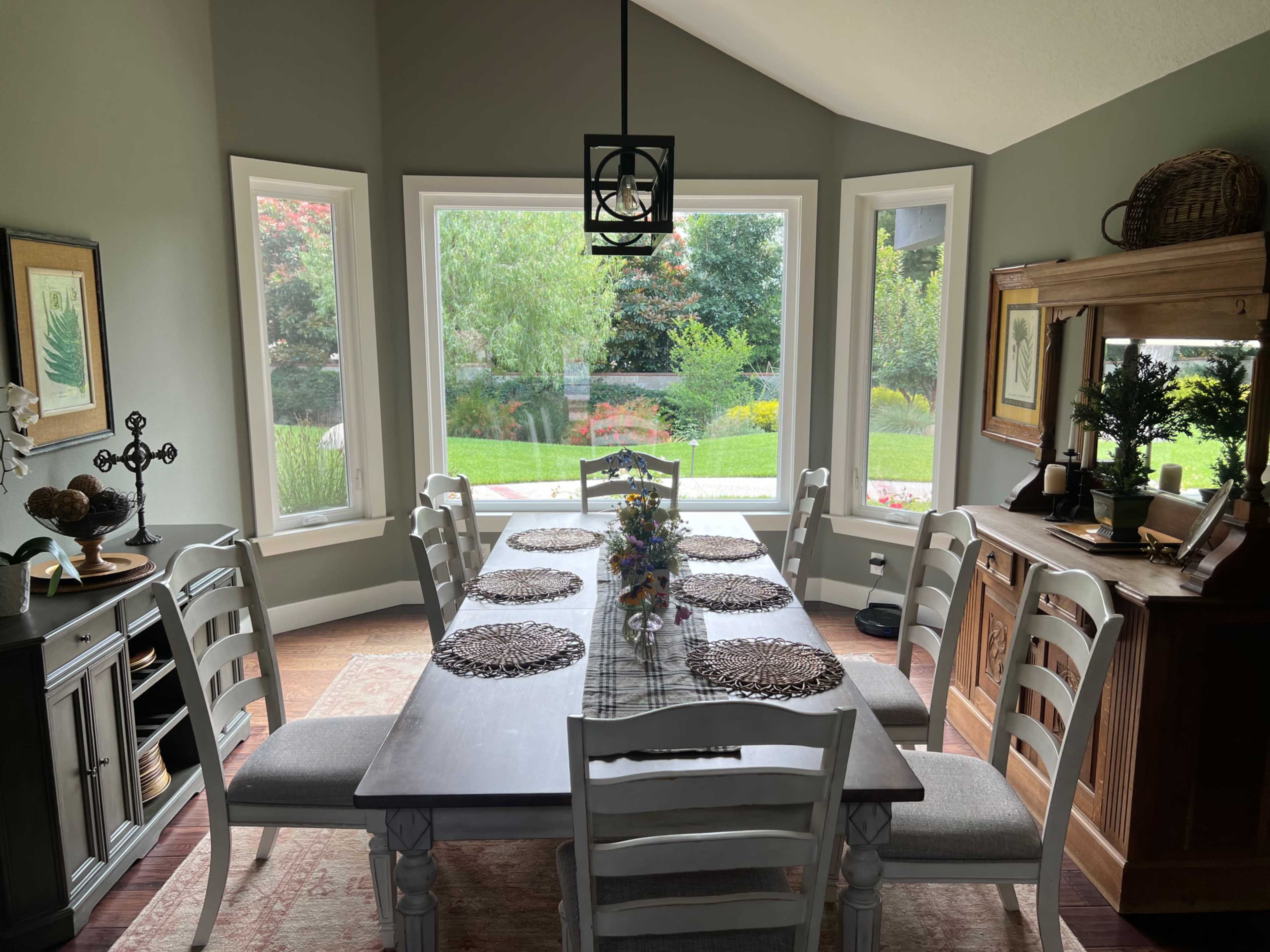 A dining room features a long wooden table set with decorative placemats and surrounded by white chairs, with large windows showcasing a garden view.