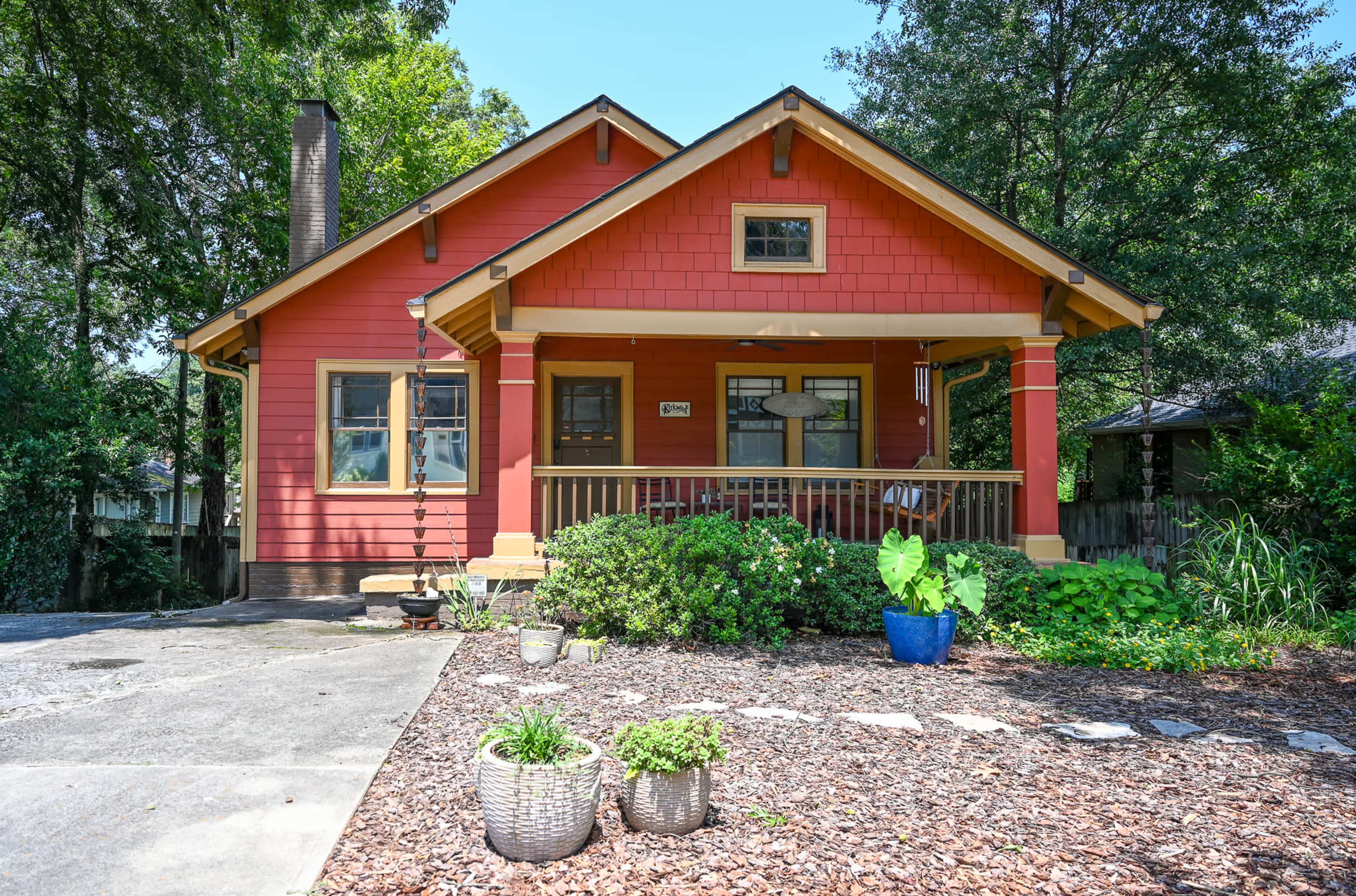 A red and tan bungalow-style house with a front porch, surrounded by landscaped greenery and decorative planters.
