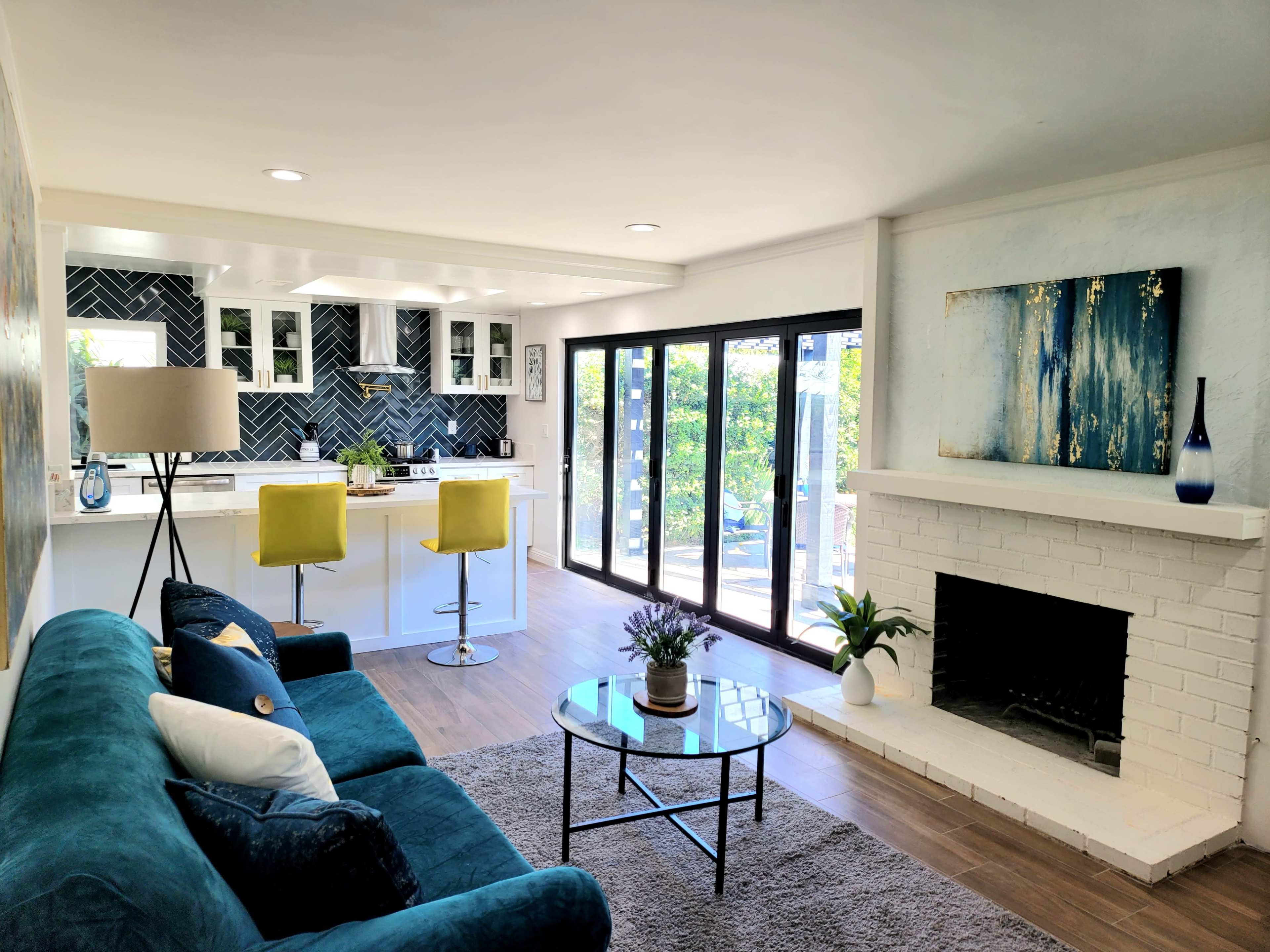 A modern living room with a teal sofa, a round coffee table, and a view of a kitchen area featuring white cabinets and a black backsplash, with sliding glass doors leading to an outdoor space.