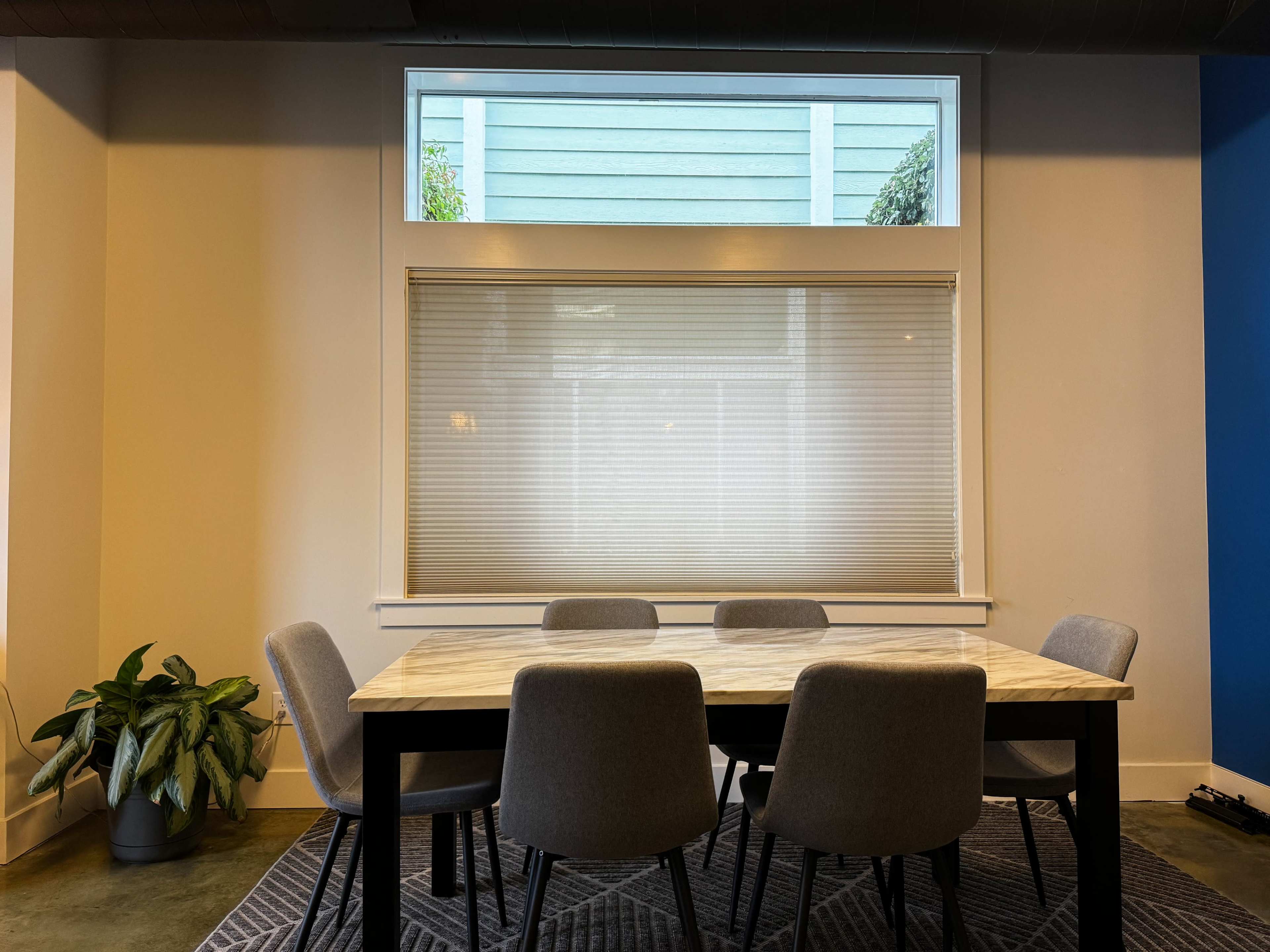 A dining table with six gray chairs is positioned in front of a large window covered with a light blind, illuminating a room with a blue accent wall.