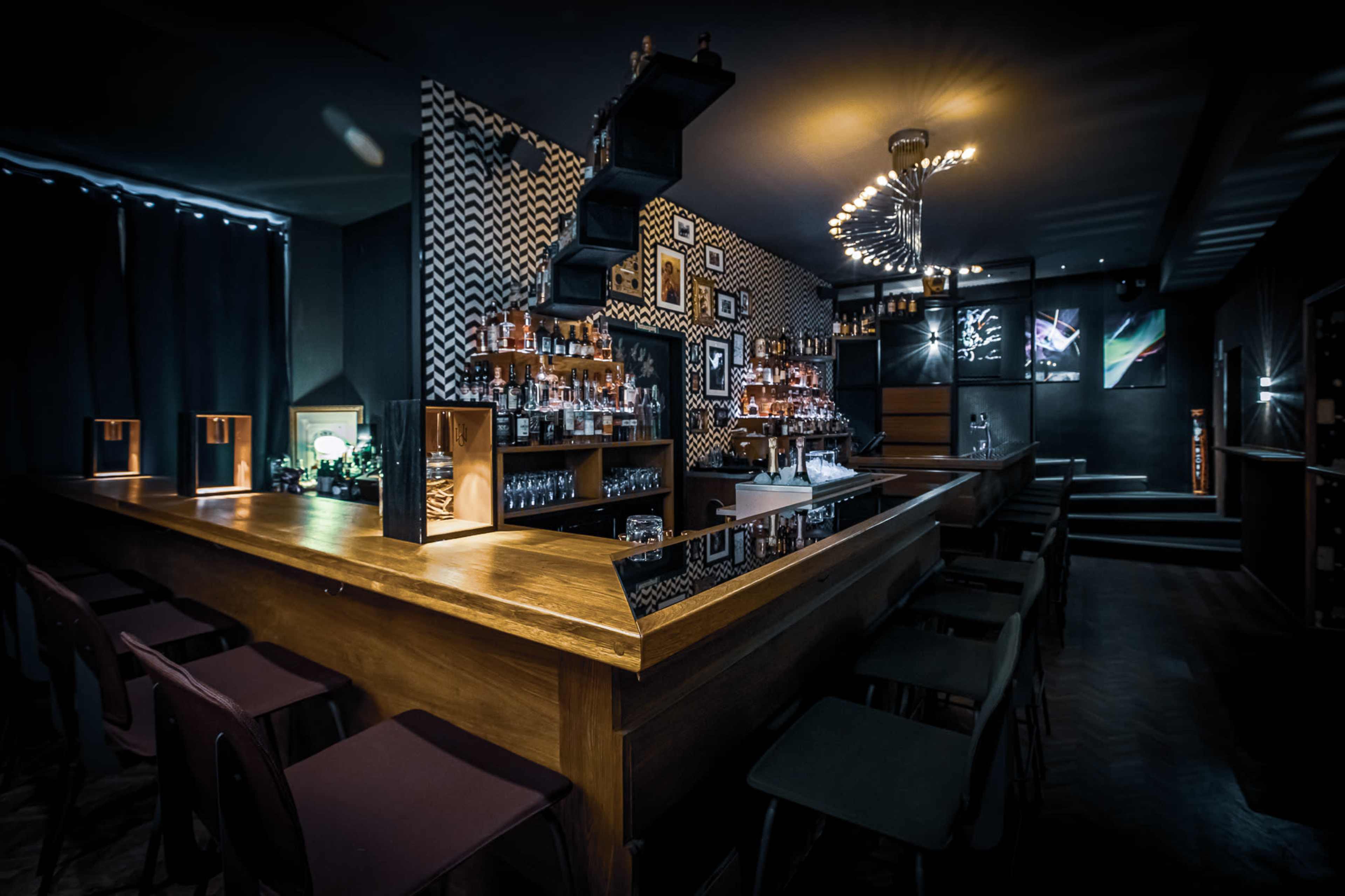 The image shows a stylish bar area with a wooden counter, high stools, and a wall decorated with geometric patterns and shelves of liquor.