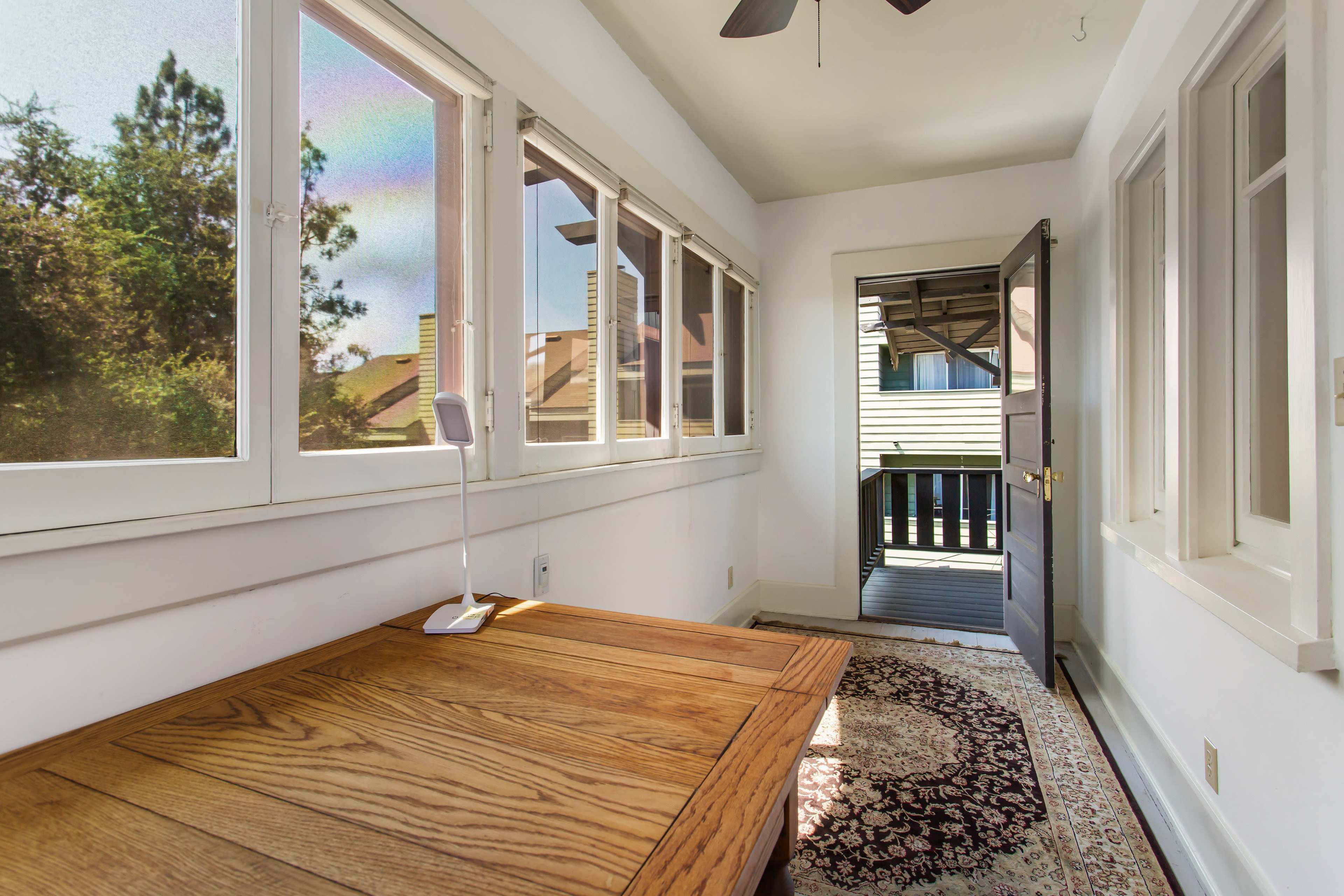 A small, bright room features a wooden table, a lamp, and large windows overlooking a balcony.