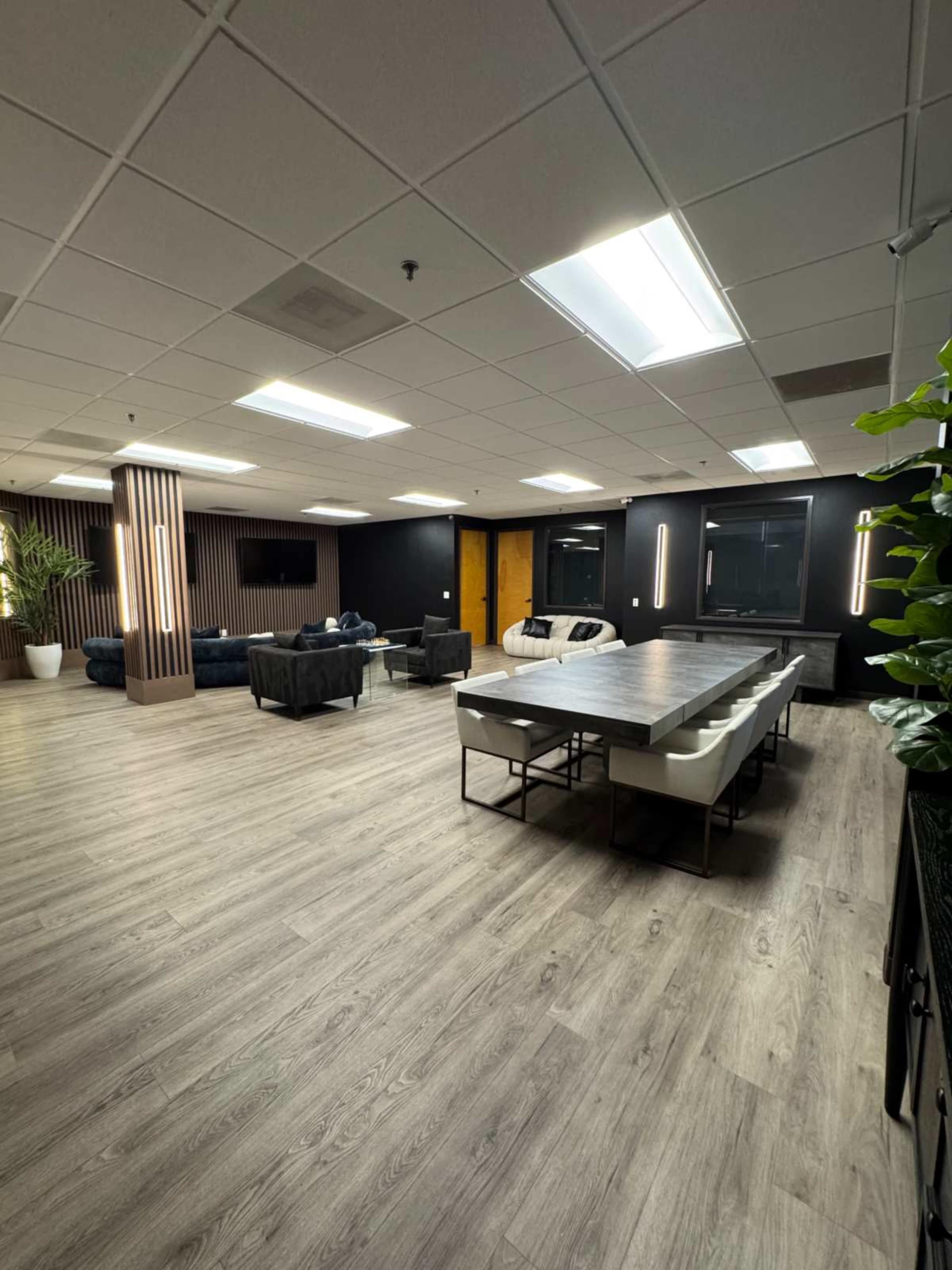 The image shows a modern office space featuring a large table and a seating area with sofas, surrounded by artificial plants and minimalist decor.