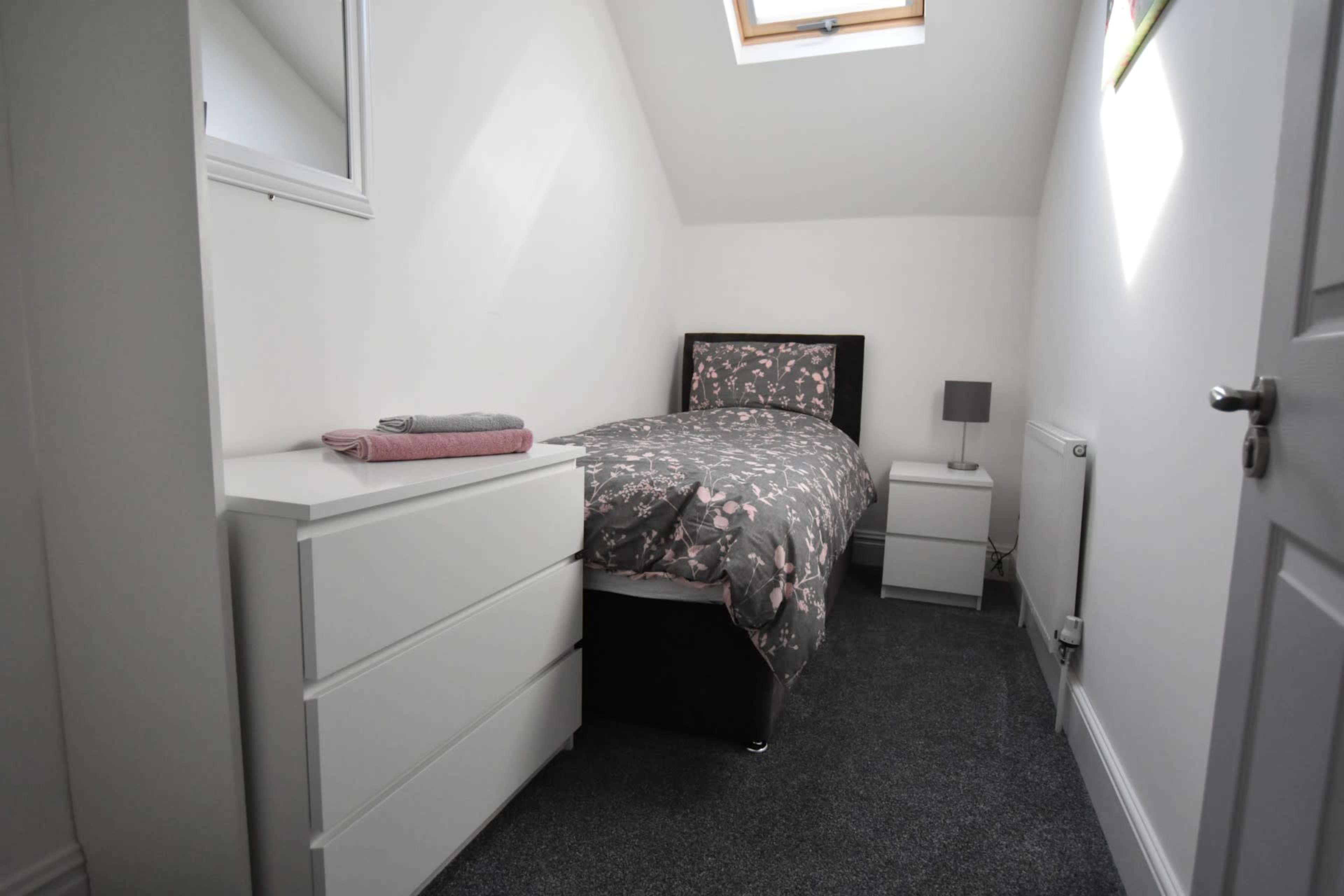 A small bedroom features a single bed with patterned bedding, a nightstand, a dresser, and a skylight.