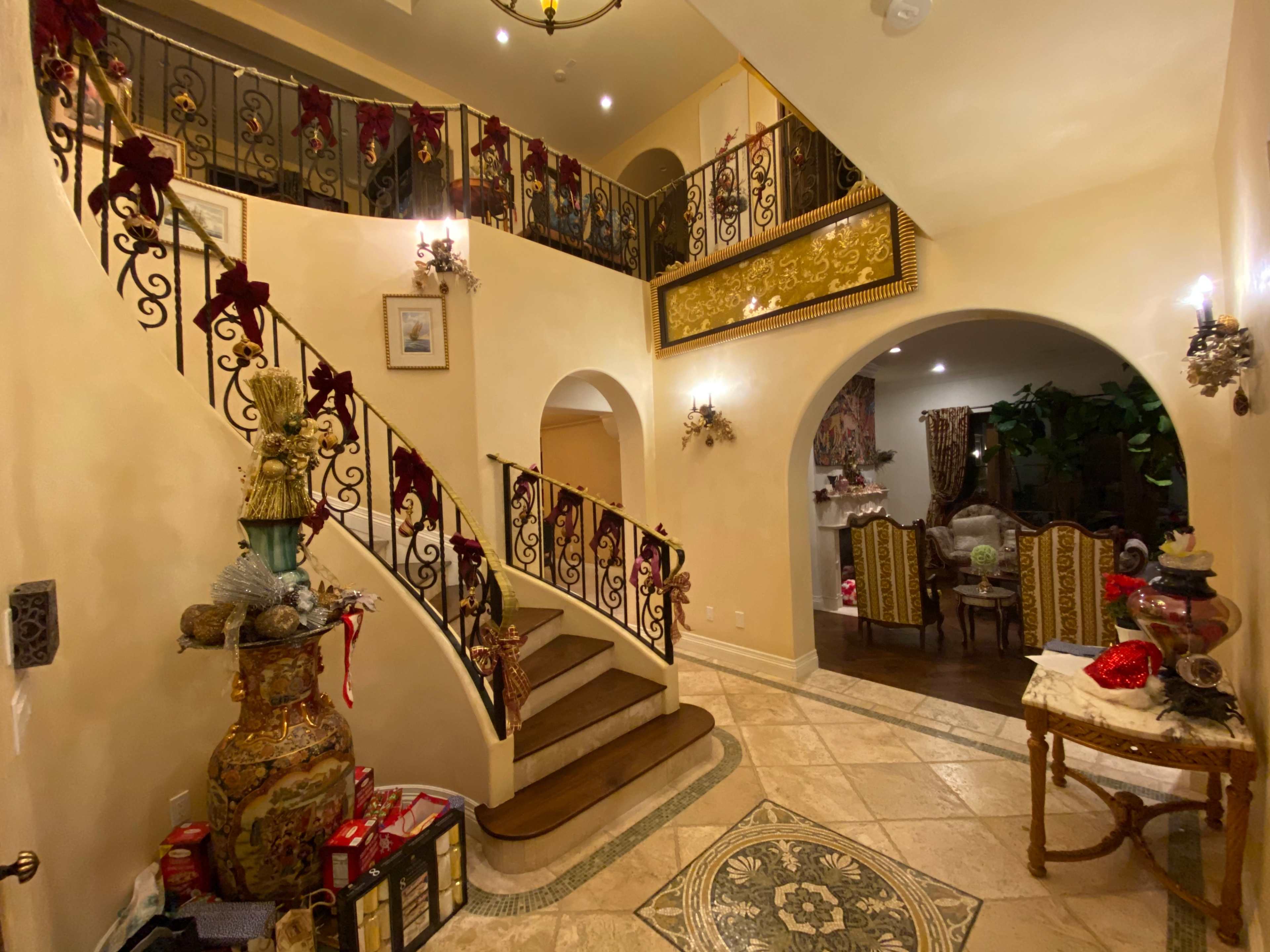 The image shows a spacious entryway featuring a staircase with ornate iron railings, decorated for the holidays, alongside a decorative table and a dining area in the background.