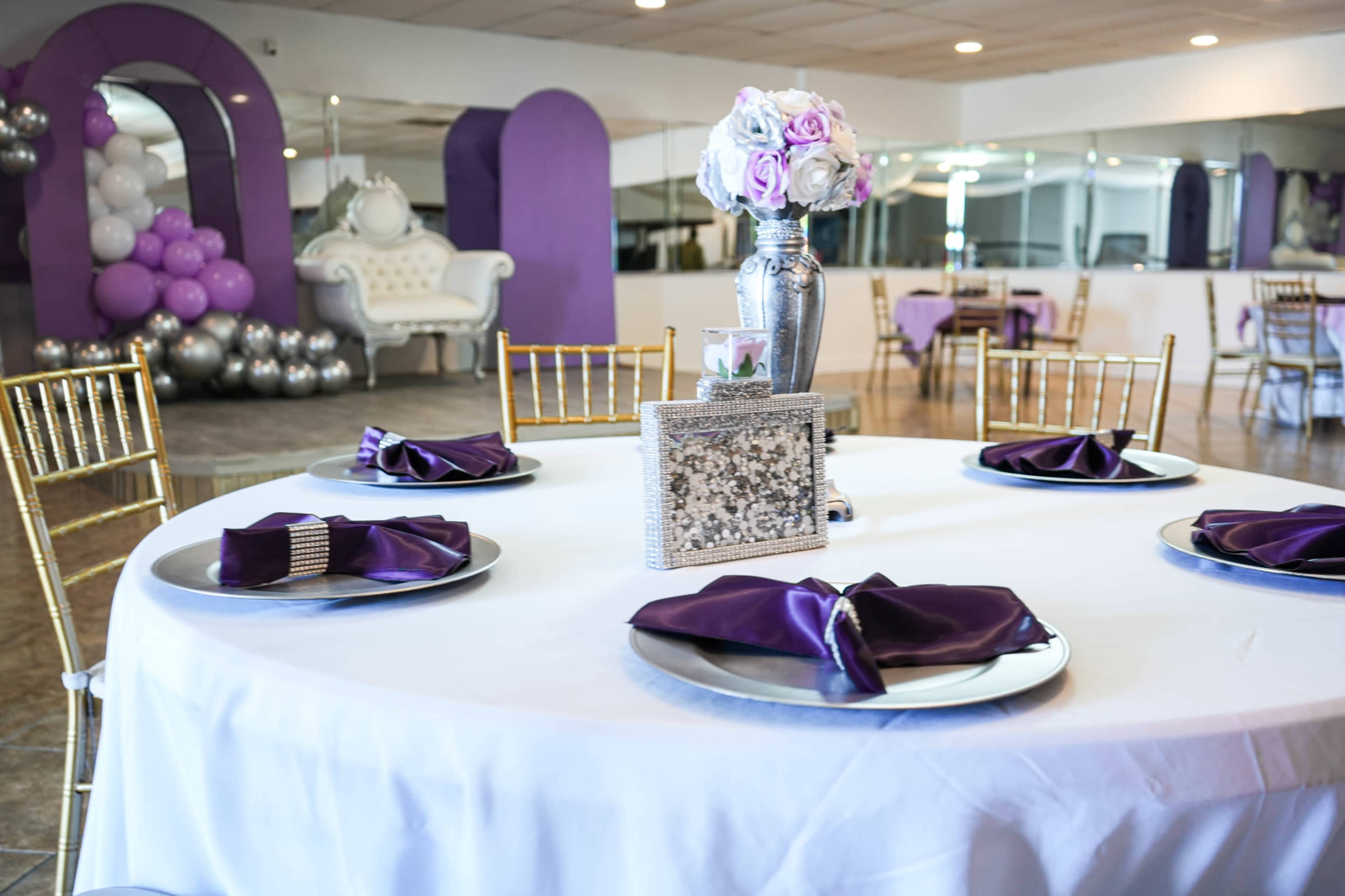 A round table is set with purple napkins and silver plates in a decorated event space featuring balloons and floral arrangements.