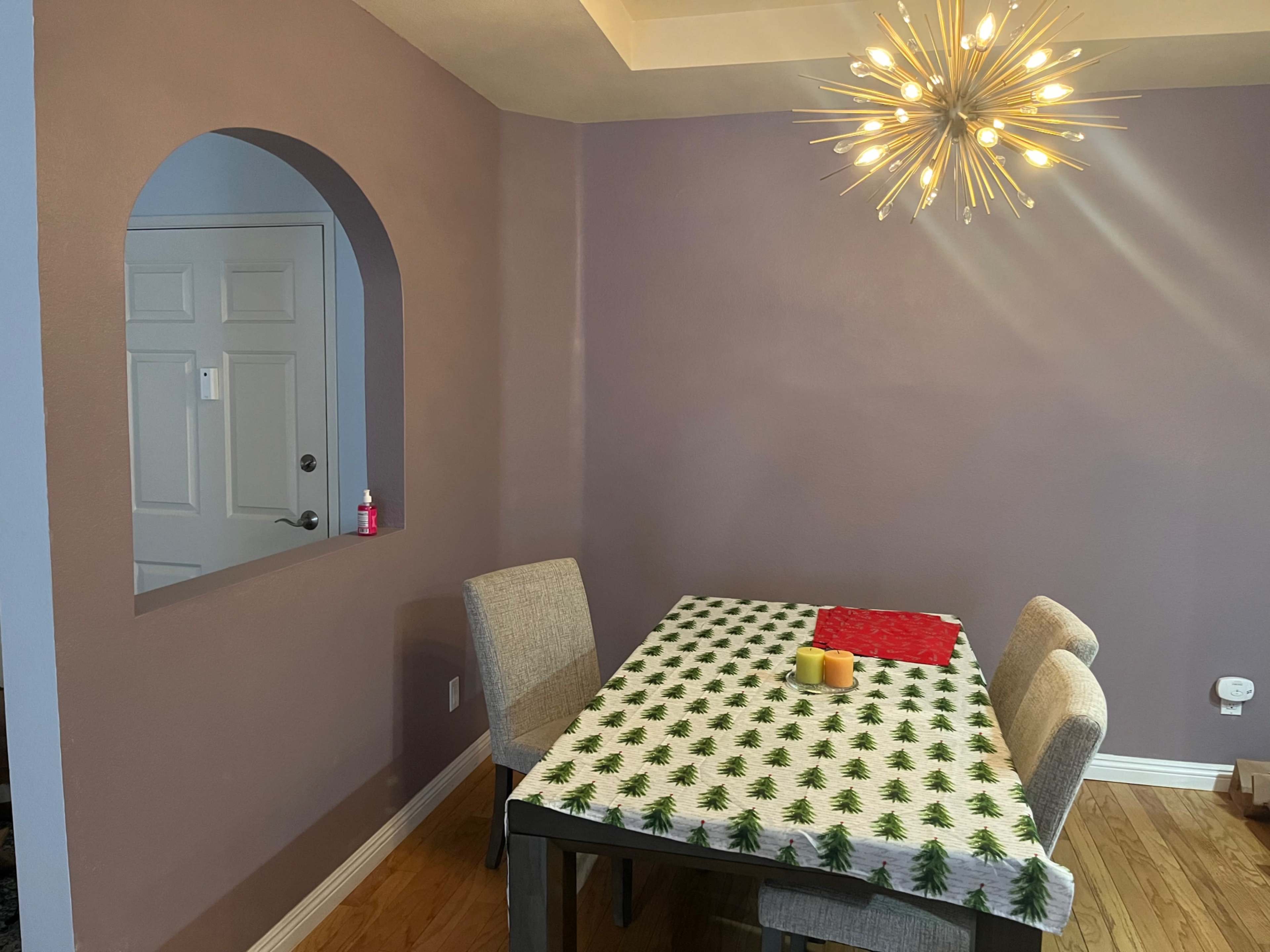 A dining area with a table covered by a holiday-themed tablecloth, four chairs, and a wall opening leading to a small area with a door.