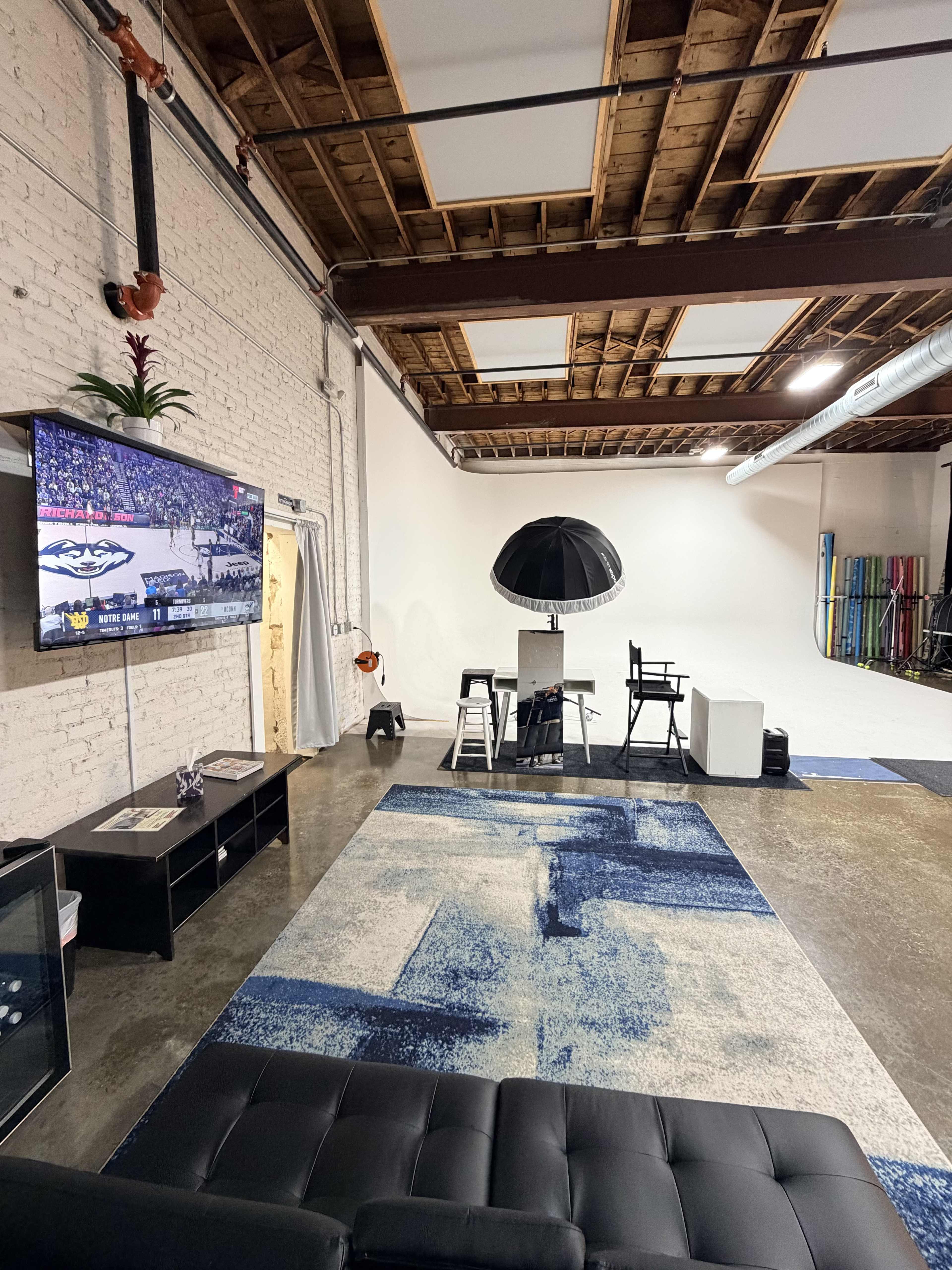 The image shows a spacious studio with a television displaying a sports game, a black sofa, a patterned rug, and various photography equipment arranged around a bright white wall.