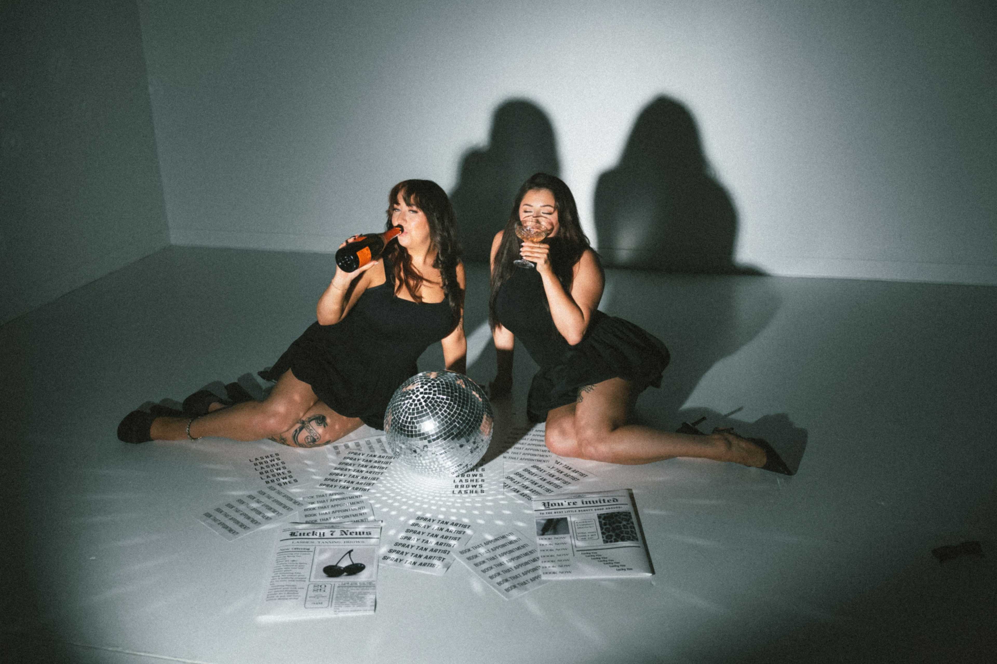 Two women sit on the floor beside a disco ball, surrounded by newspaper articles and drink bottles, in a well-lit studio setting.