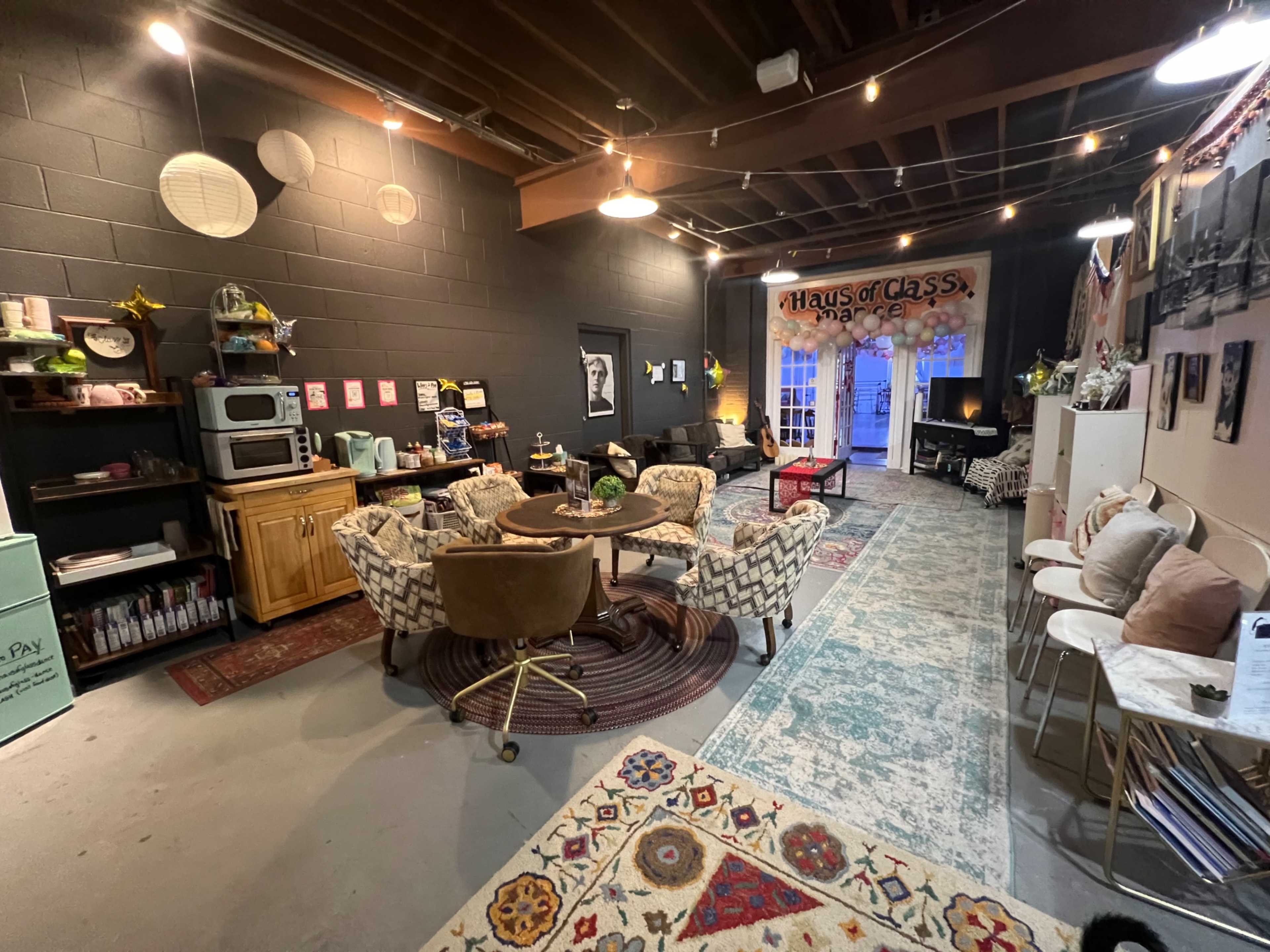 A cozy interior space with a mix of seating arrangements, decorative rugs, and bright lighting, featuring a kitchen area and artwork on the walls.
