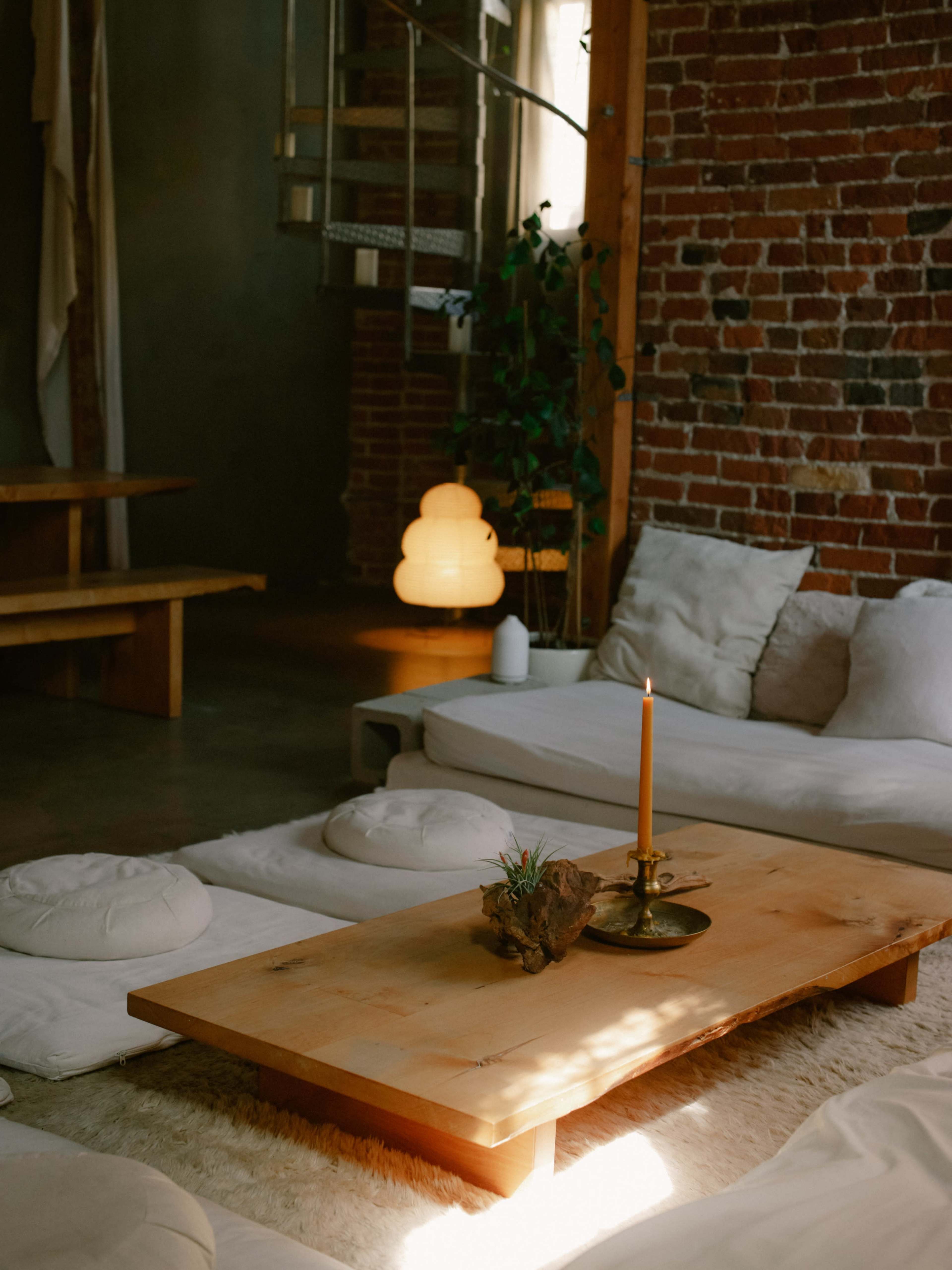 The scene shows a cozy, minimalist living area featuring a wooden coffee table with a candle, surrounded by soft seating cushions on a light rug, and warm lighting from a nearby lamp.