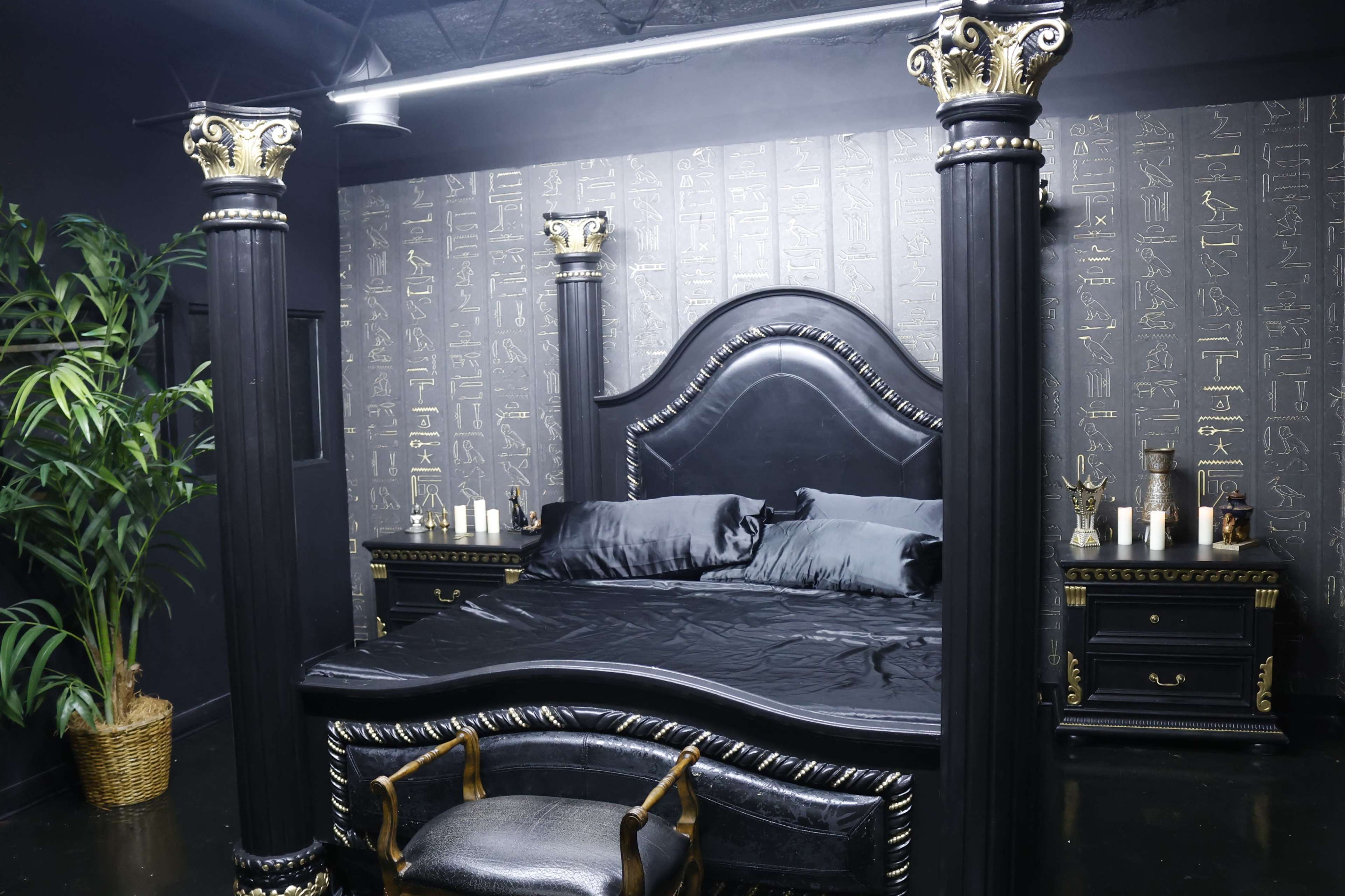 A dark-themed bedroom with a large, ornate black bed featuring tall, gilded columns, surrounded by stylish furniture and a decorative plant.