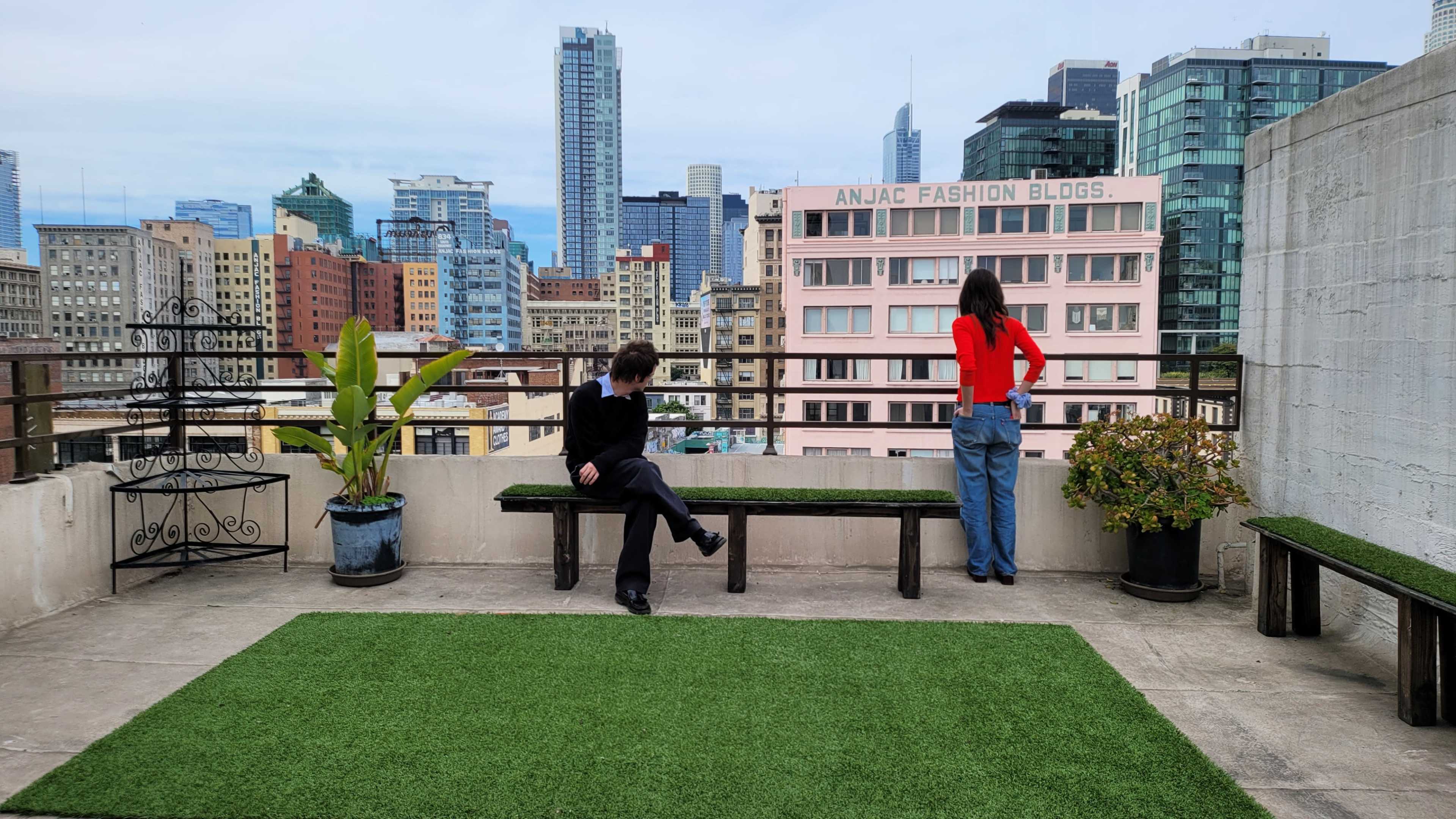 Two people stand on a rooftop terrace adorned with potted plants, overlooking a city skyline filled with high-rise buildings.