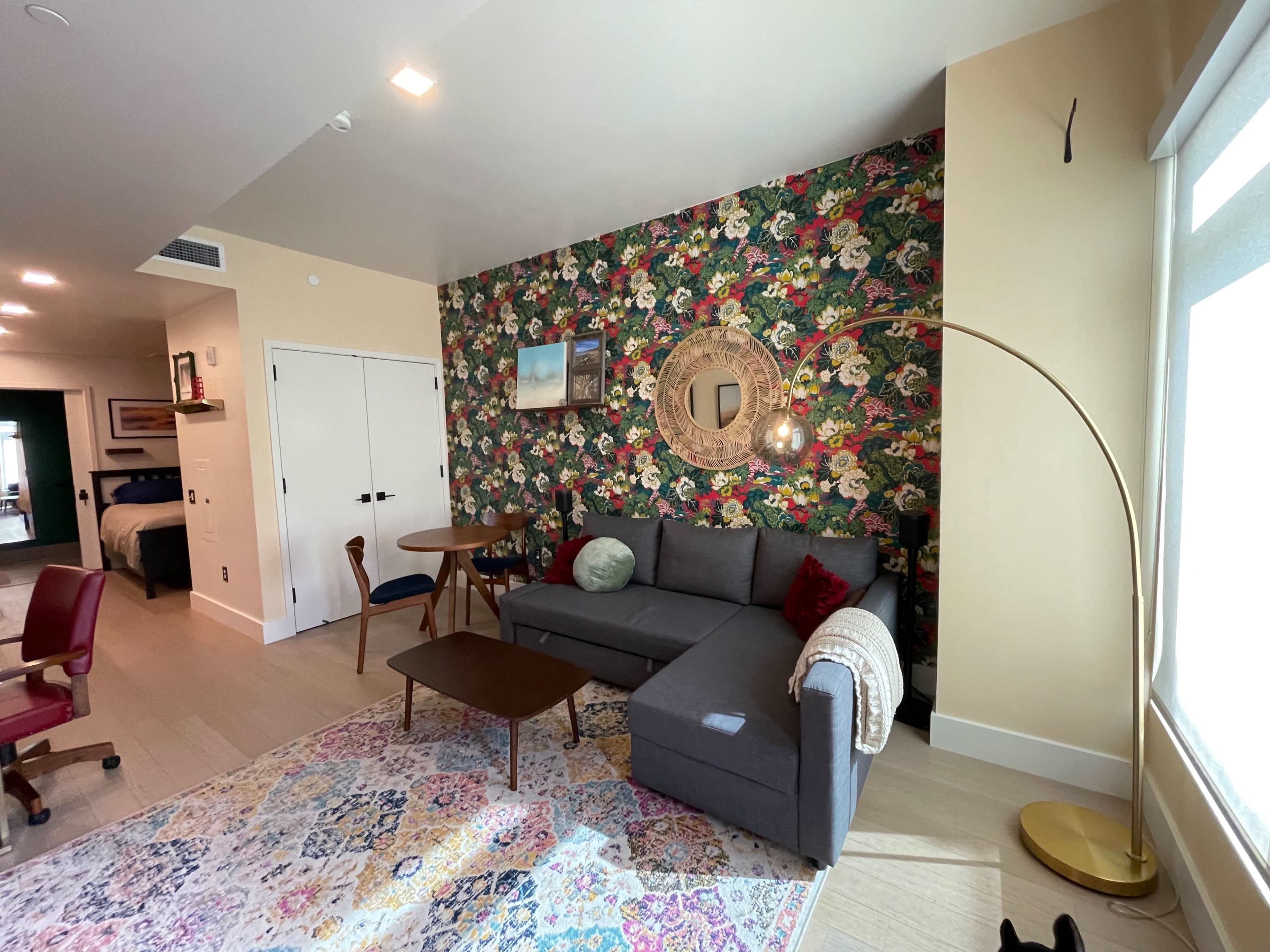 A living room featuring a floral accent wall, a gray sectional sofa, a round mirror, and a floor lamp beside a small table and chairs.
