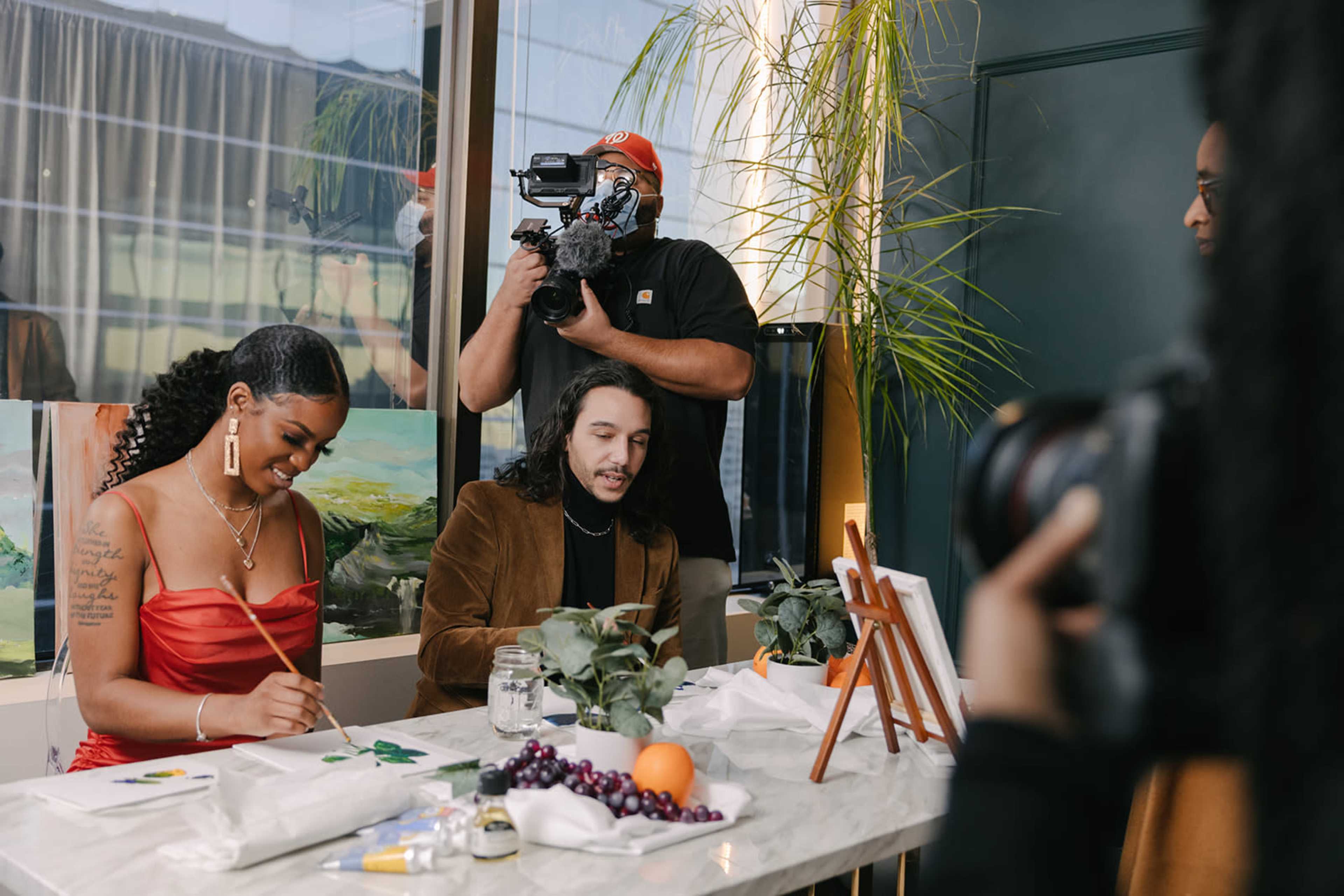 A woman in a red dress paints at a table while a man in a brown jacket writes, as a cameraman films the scene.