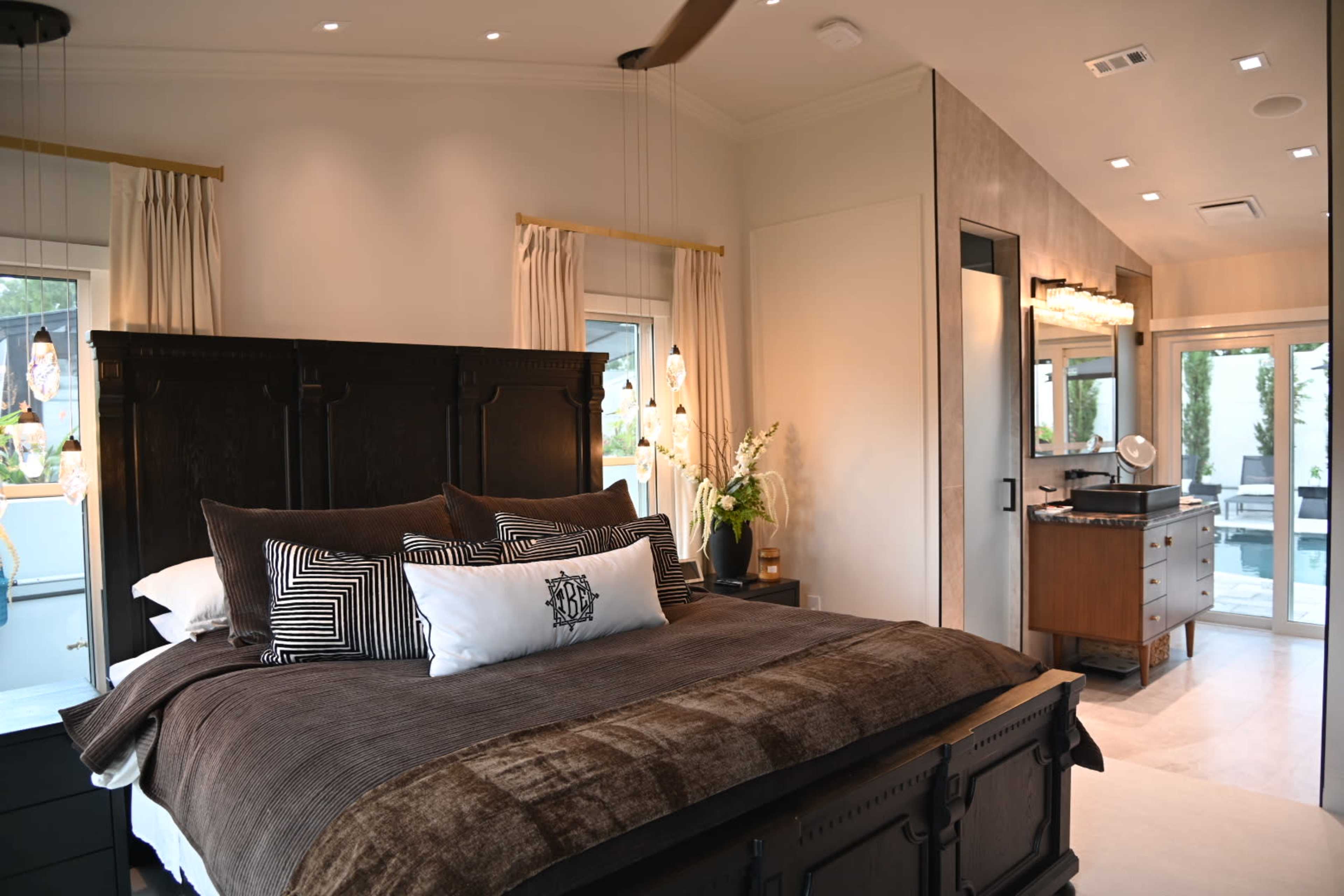 The image shows a modern bedroom featuring a large bed with dark wooden furnishings, decorative pillows, and large windows that provide natural light, alongside a sleek bathroom area.