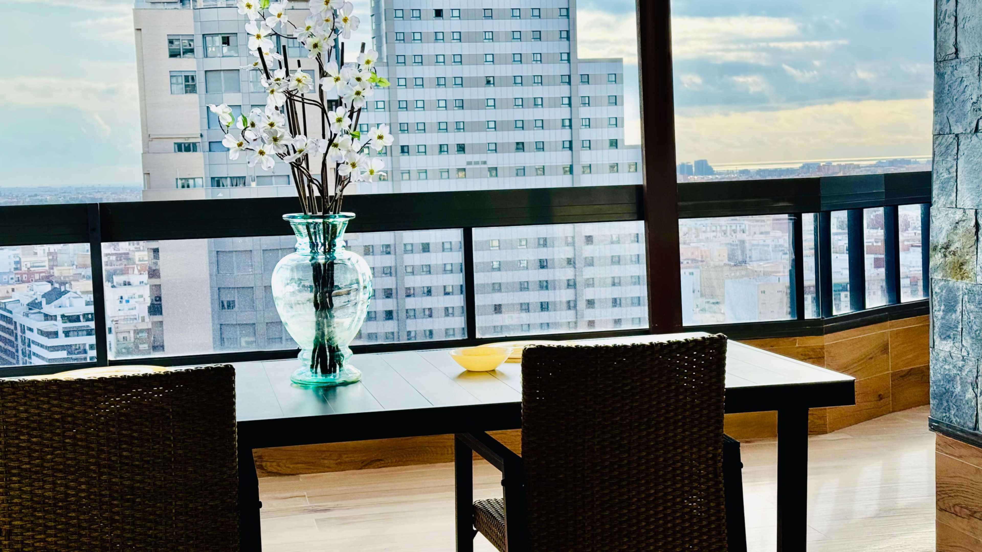 A dining table with two chairs and a vase of flowers overlooks a cityscape from a high-rise building.