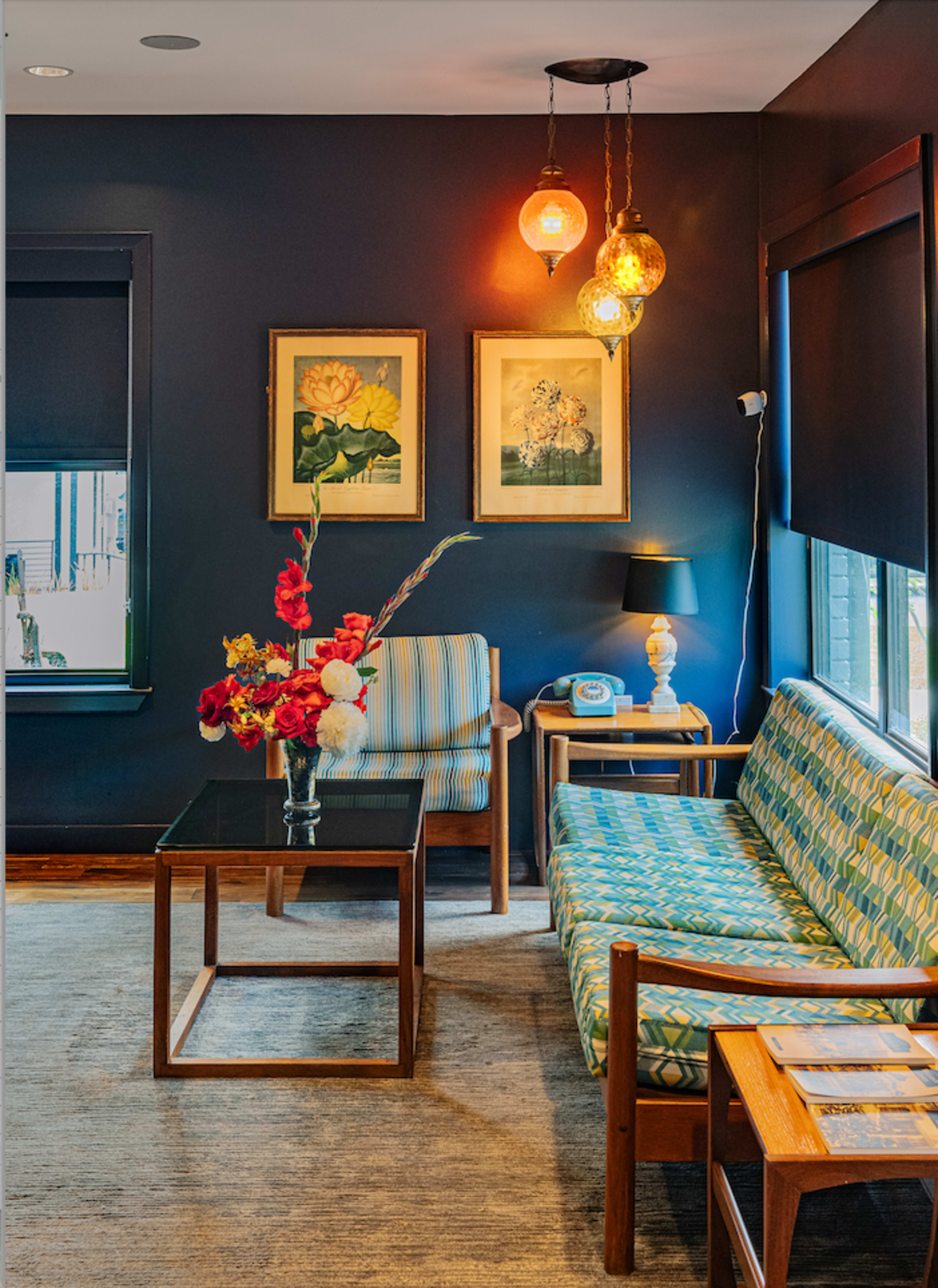 The image depicts a cozy living room featuring a patterned sofa, a glass coffee table, floral arrangements, and framed artwork on the dark-colored walls.
