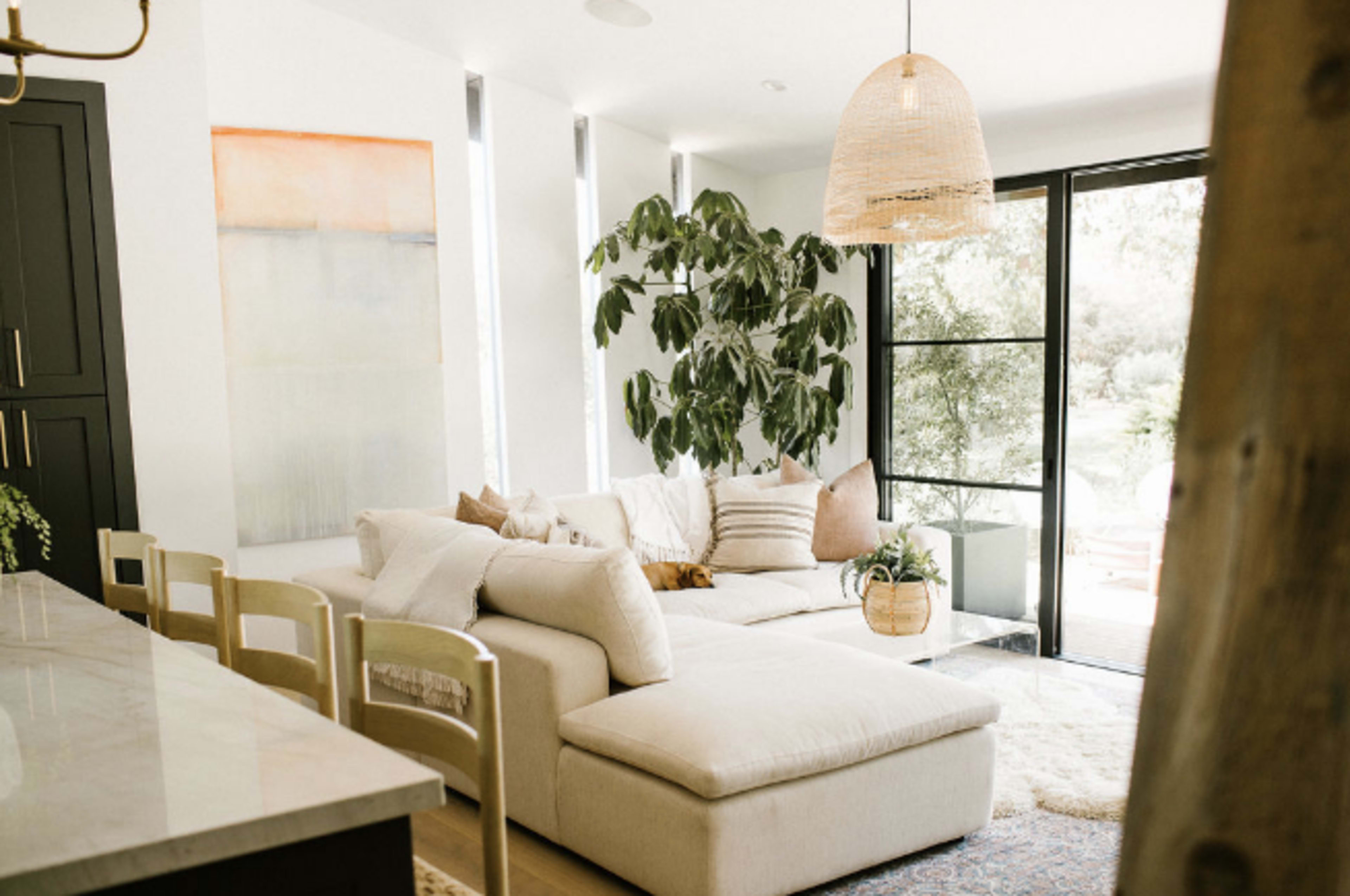 The image shows a bright living room with a light-colored sectional sofa, a large indoor plant, and a woven pendant light, featuring large windows that open to an outdoor space.