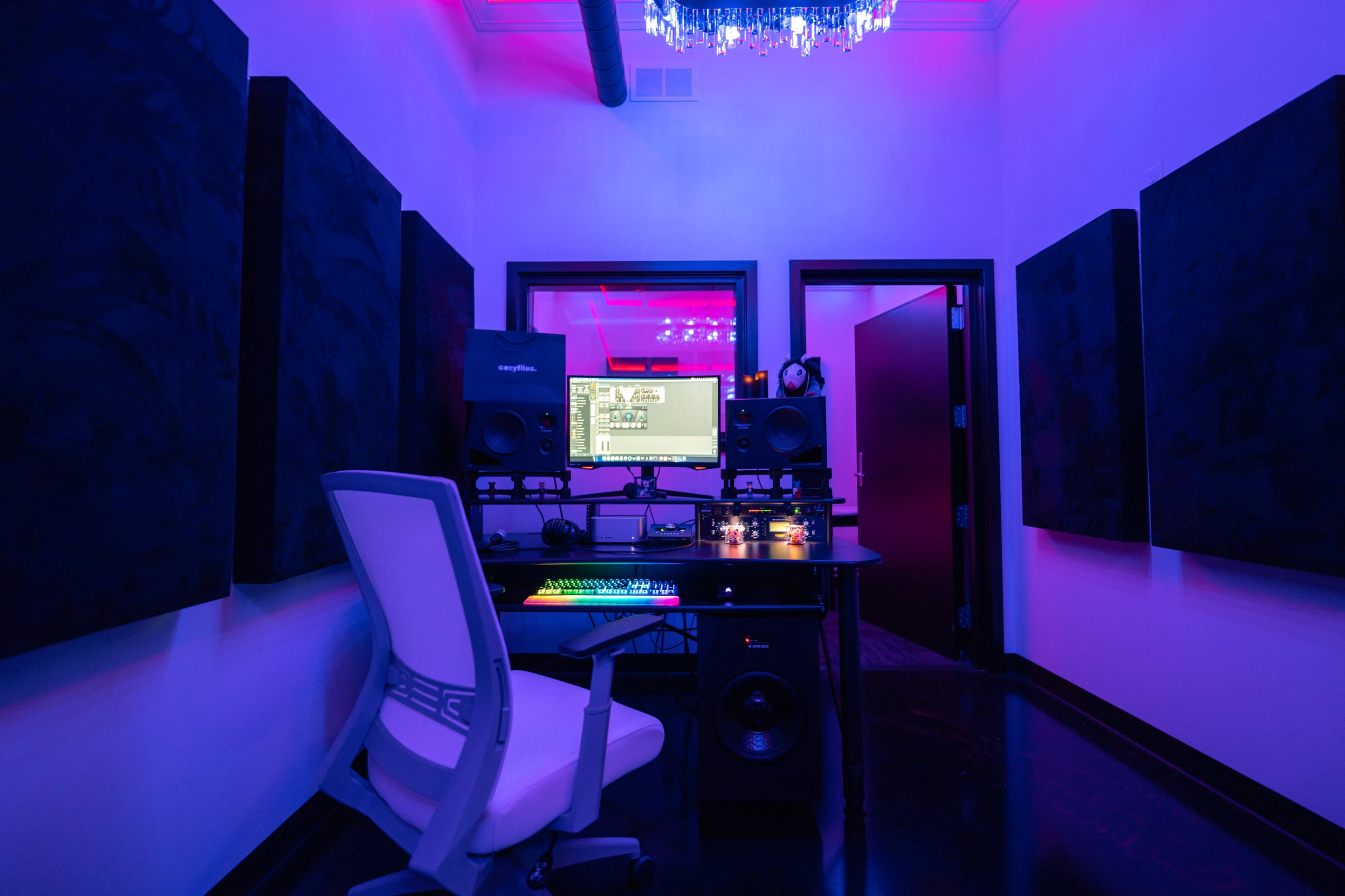 The image shows a recording studio with a desk, computer, and sound equipment, surrounded by soundproof panels and illuminated by colorful lighting.