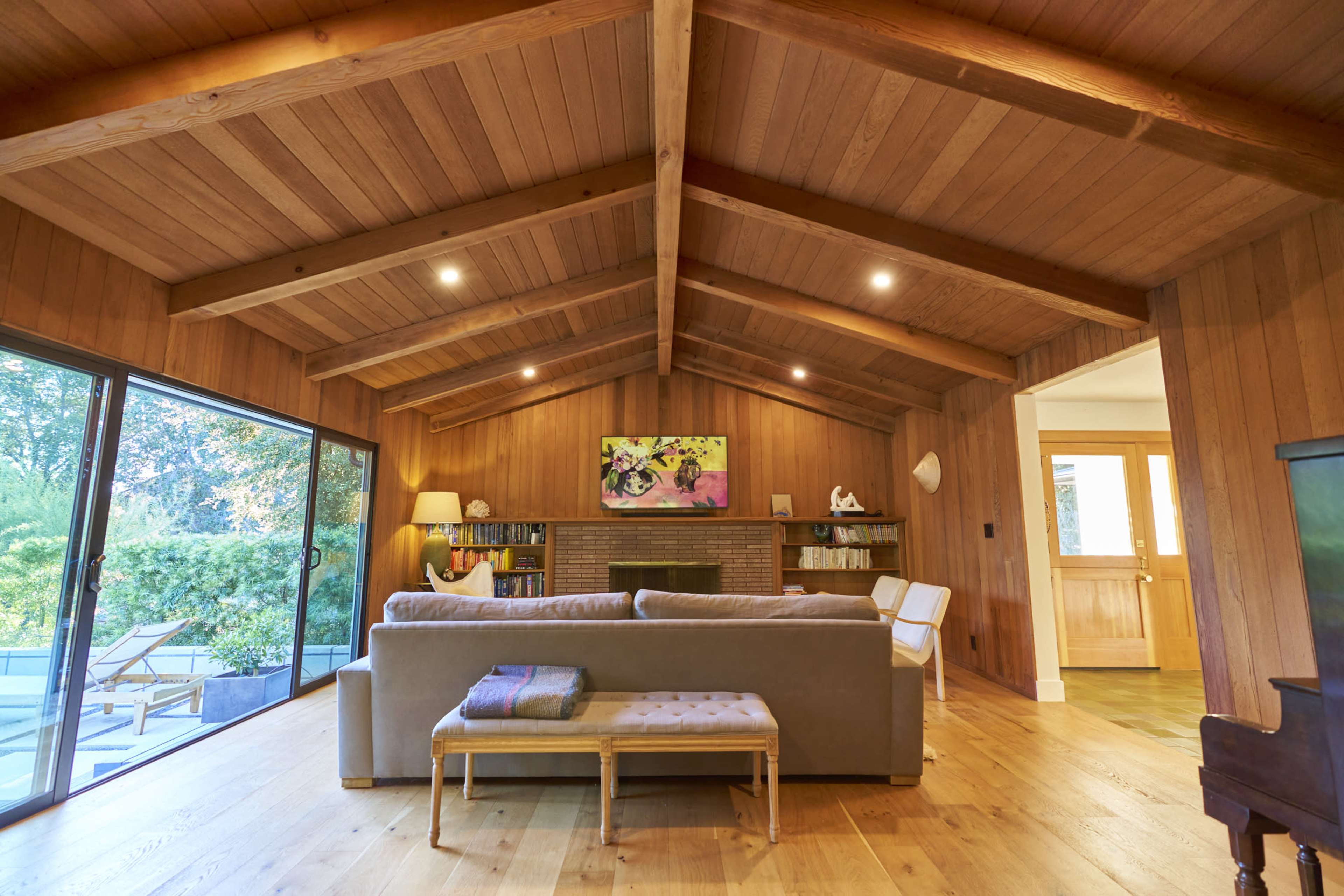 A spacious living room features wooden walls and beams, large windows overlooking a garden, and modern furnishings including a grey sofa and a light-colored armchair.