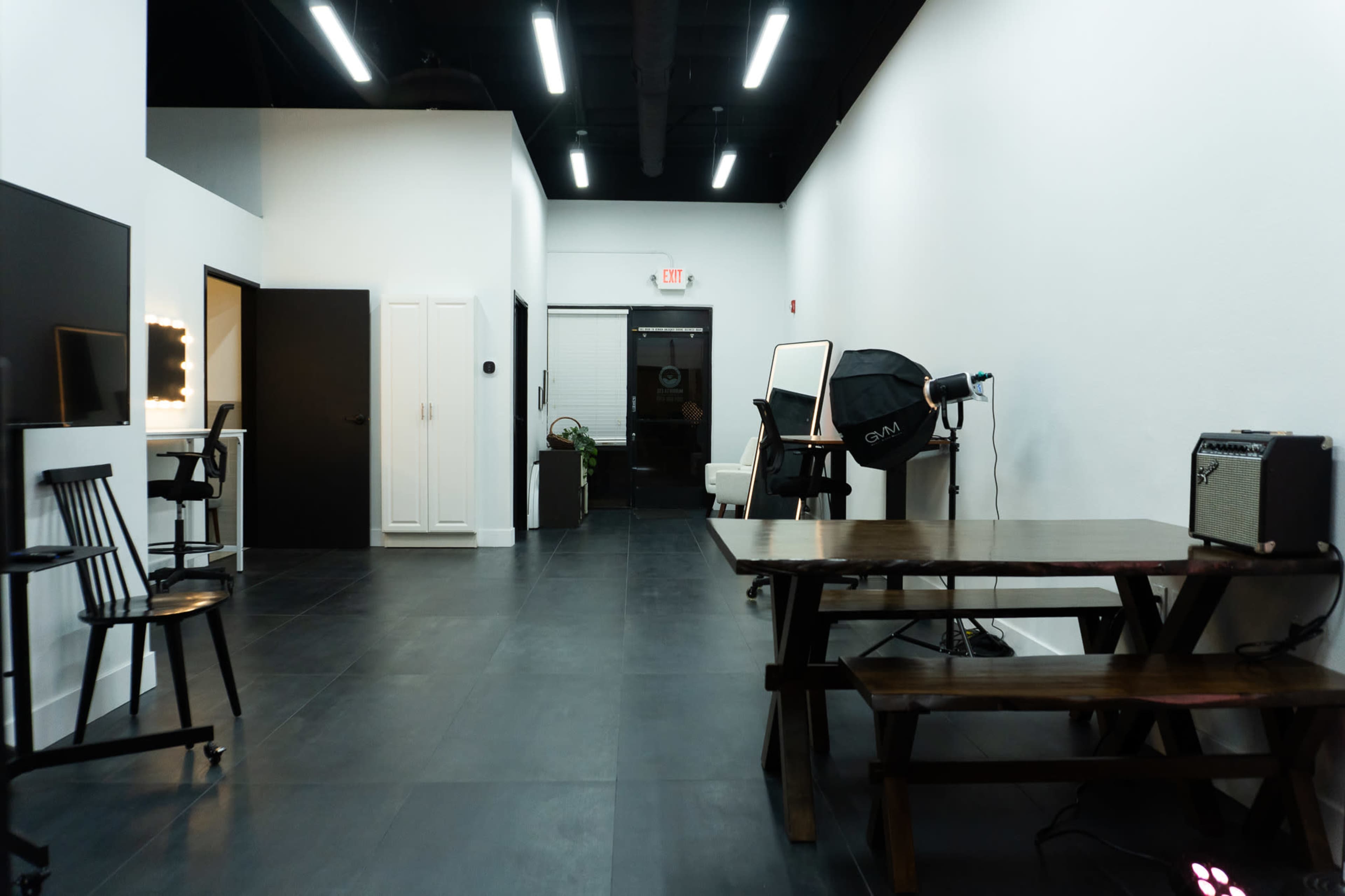 The image shows a spacious, contemporary studio interior with a black tile floor, white walls, and various furniture items, including a wooden table and chairs, a dressing mirror, and studio lighting equipment.