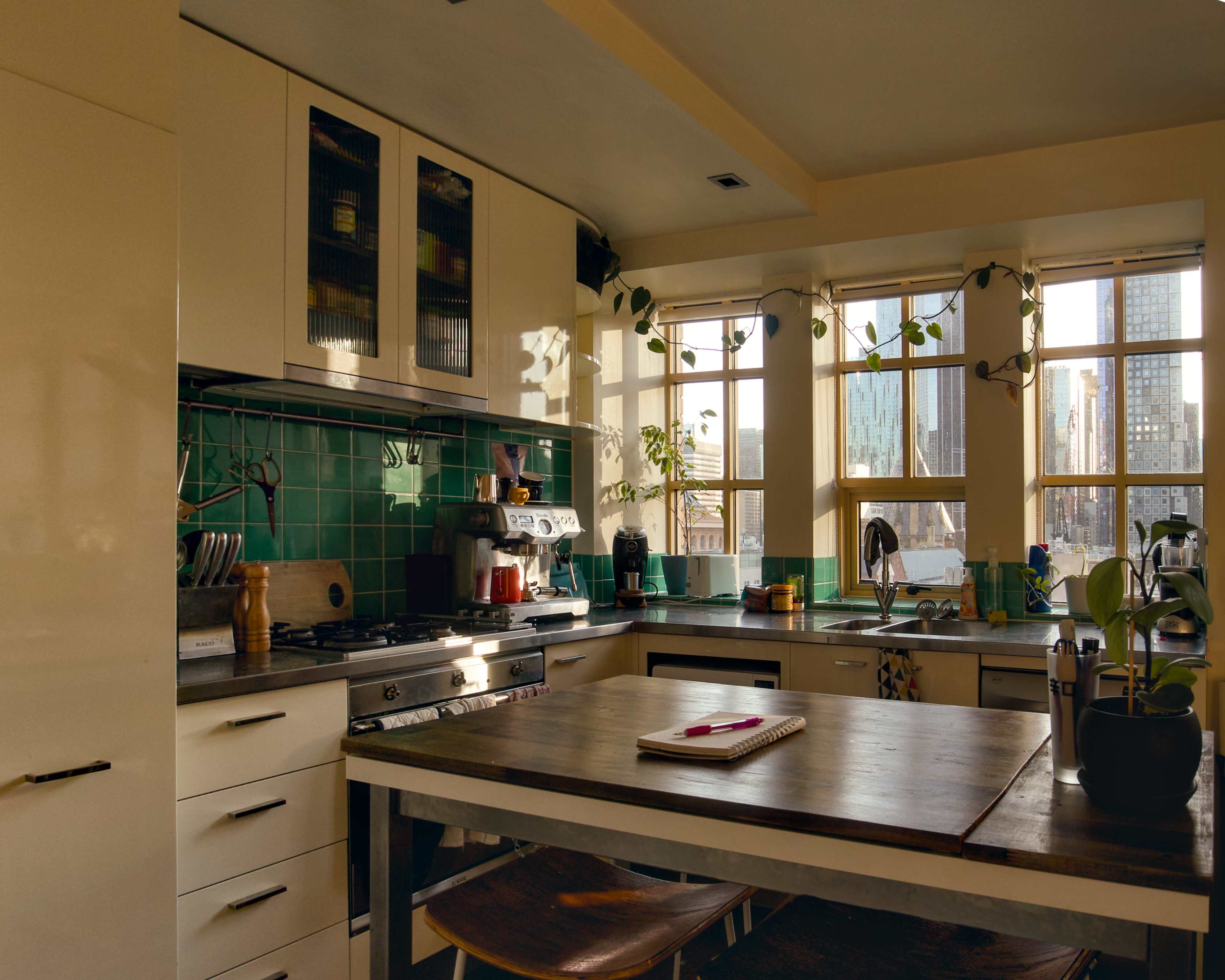 A modern kitchen with green tiled walls, a wooden table in the center, and large windows offering a view of the city skyline.