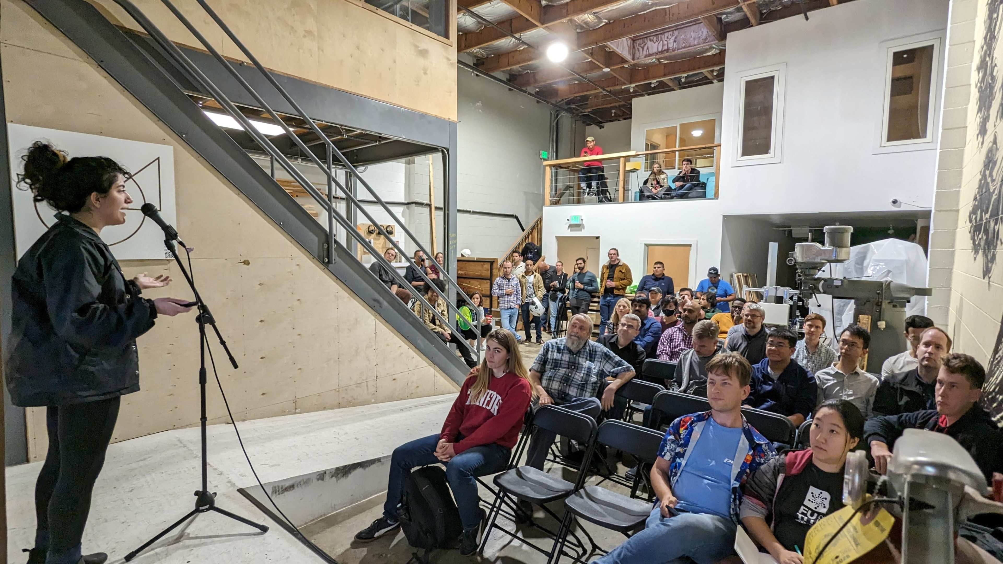 A large crowd listens attentively as a speaker addresses them from a microphone on a stage in a well-lit indoor space.