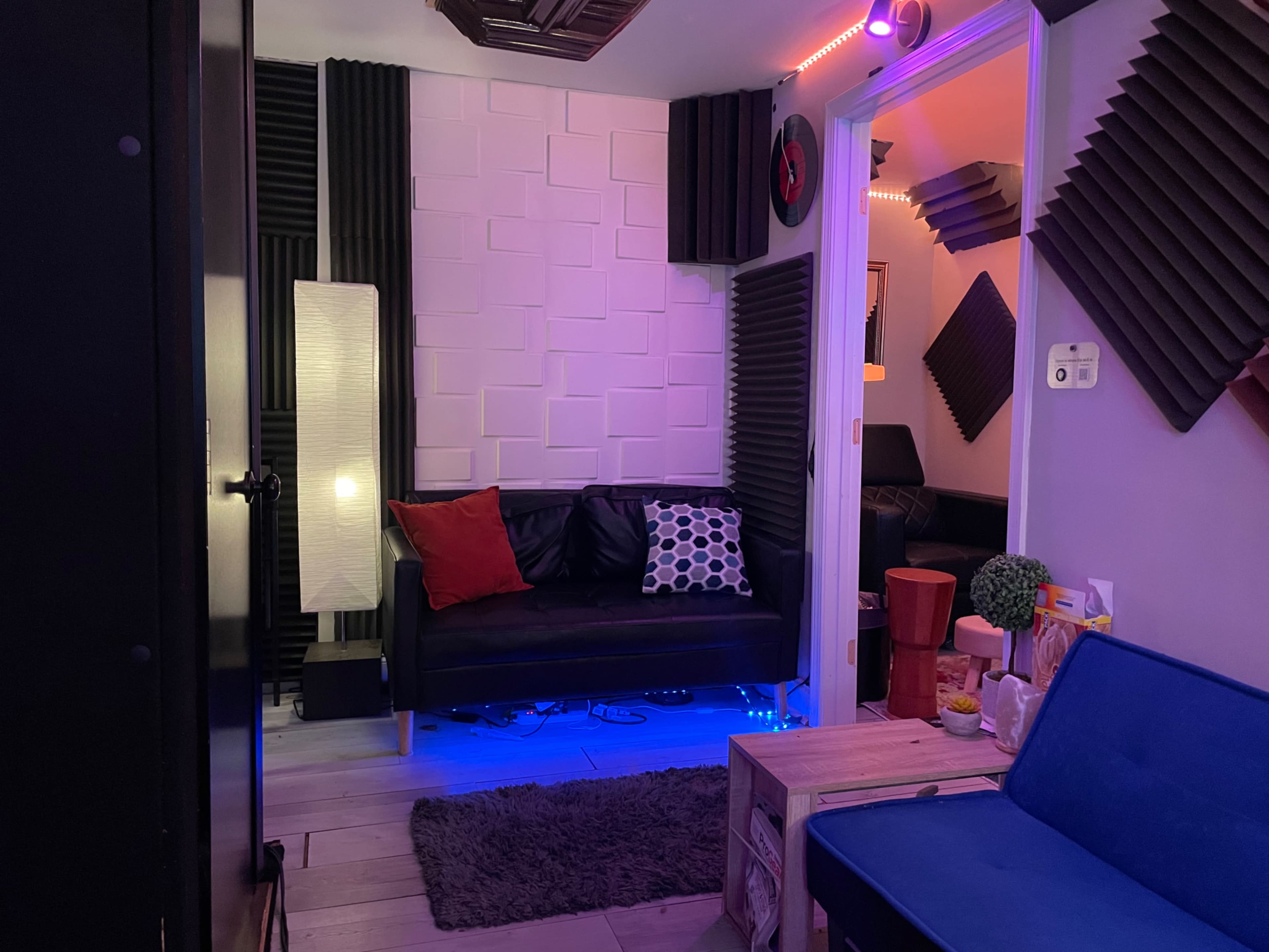 A small room with soundproofing panels on the walls, a black couch with decorative pillows, and various furnishings including a lamp and a coffee table.