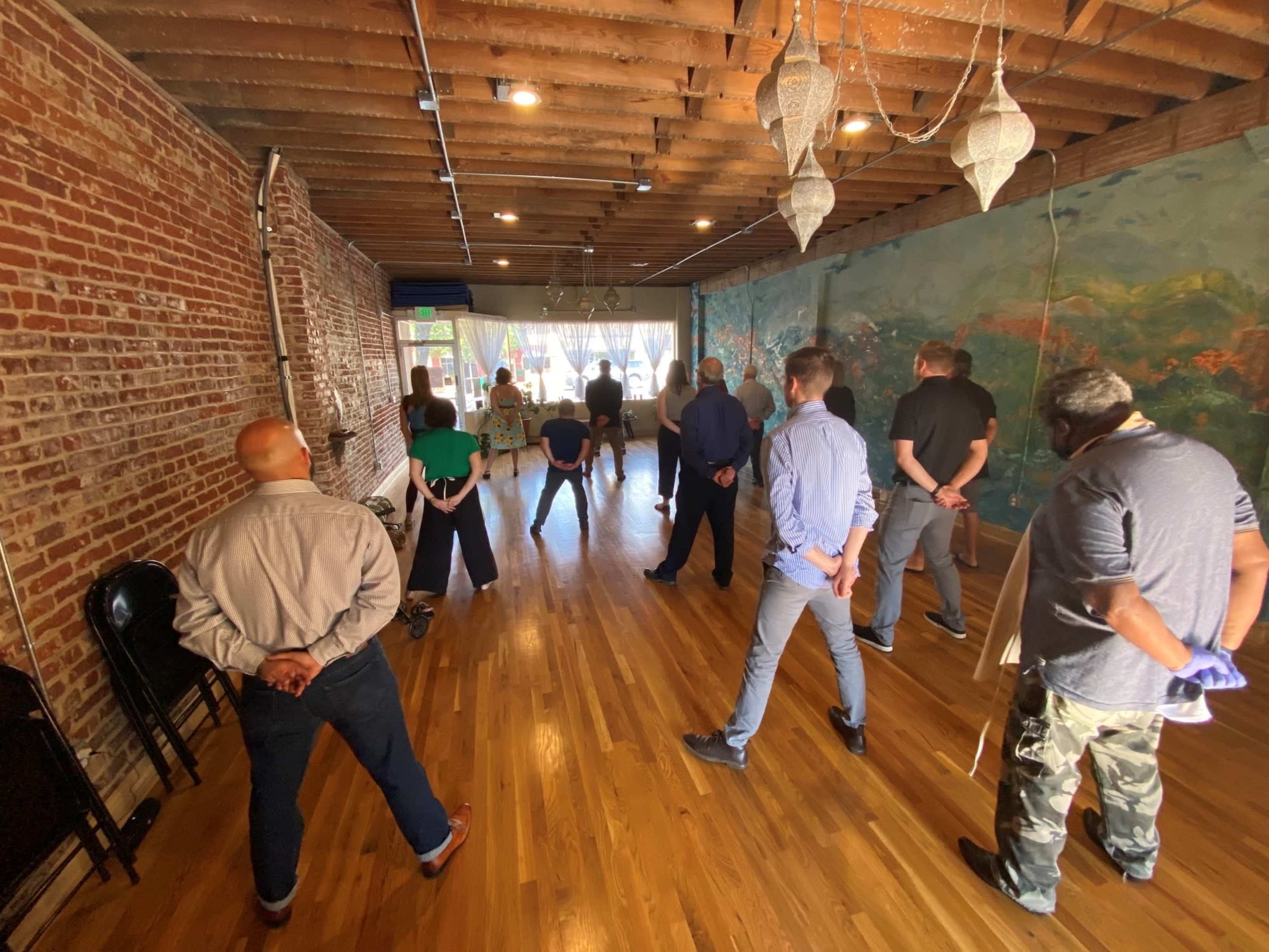 A group of people stands in a studio with exposed brick walls and a mural, facing a speaker at the front.