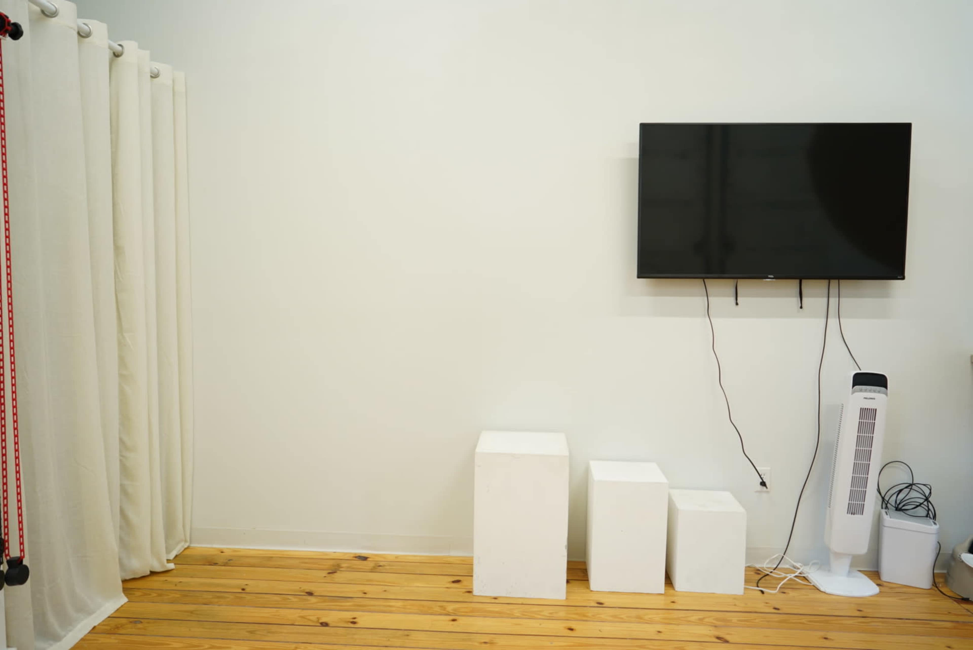 The image shows a minimalistic room with a blank wall, a mounted television, three white cubes, and a standing fan on a wooden floor.