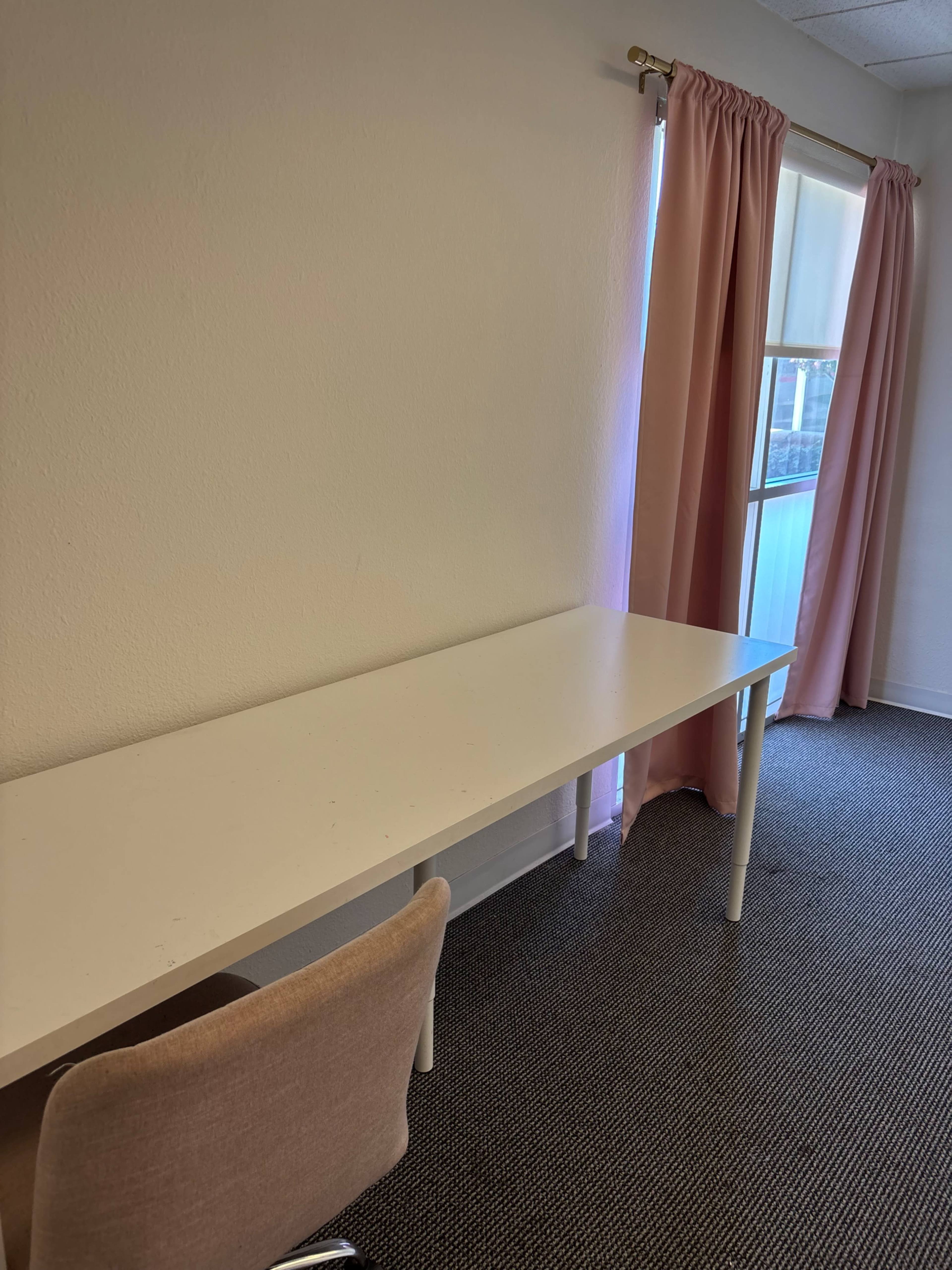 A simple white desk and a beige chair are positioned next to a window covered with pink curtains in a sparsely decorated room.