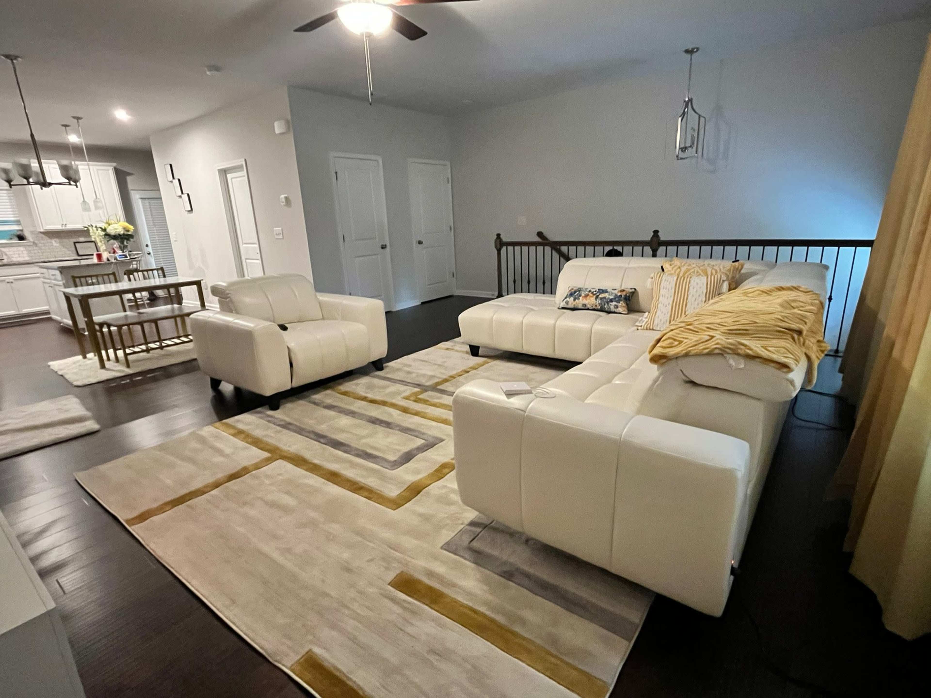 The image shows a modern living room with a white sectional sofa, a matching armchair, and a large area rug featuring geometric patterns, adjacent to a dining space with a table and chairs.