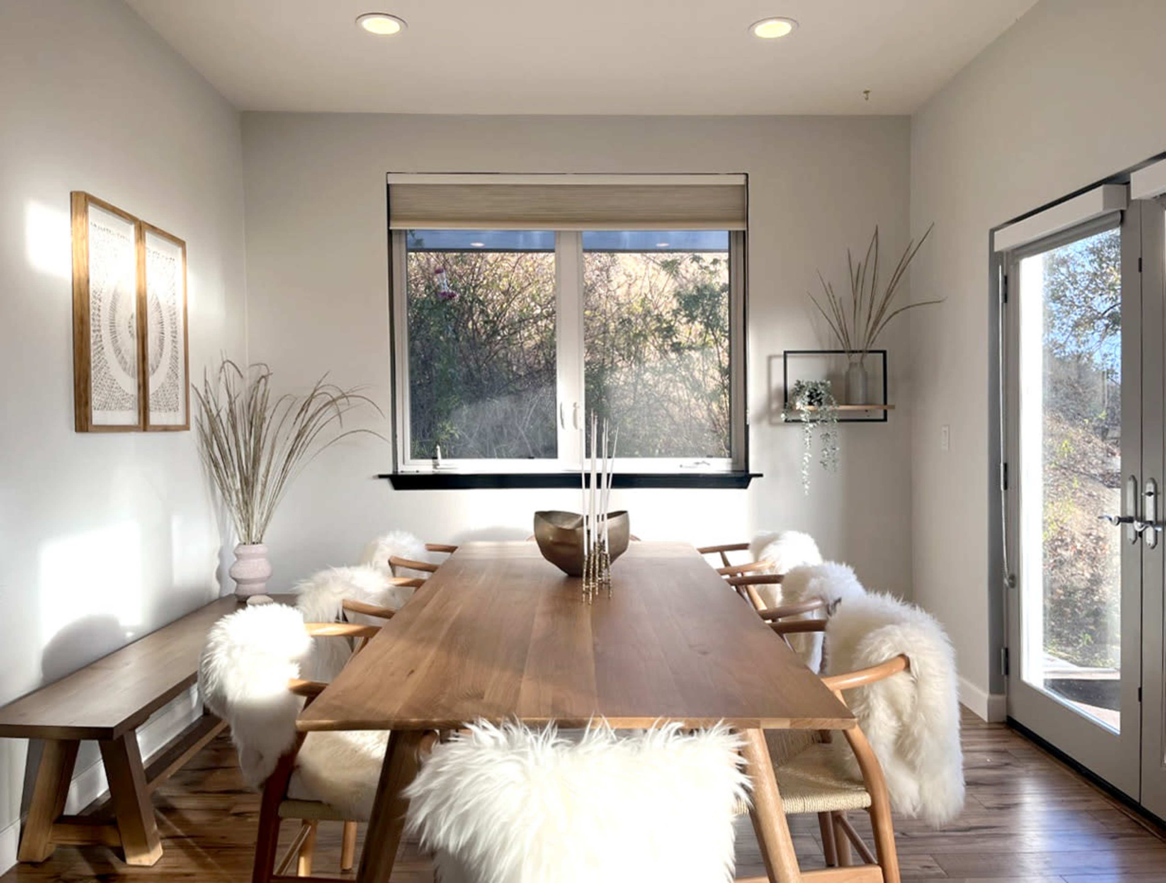 A modern dining room features a wooden table surrounded by chairs with white fur throws, complemented by large windows and minimalistic decor.