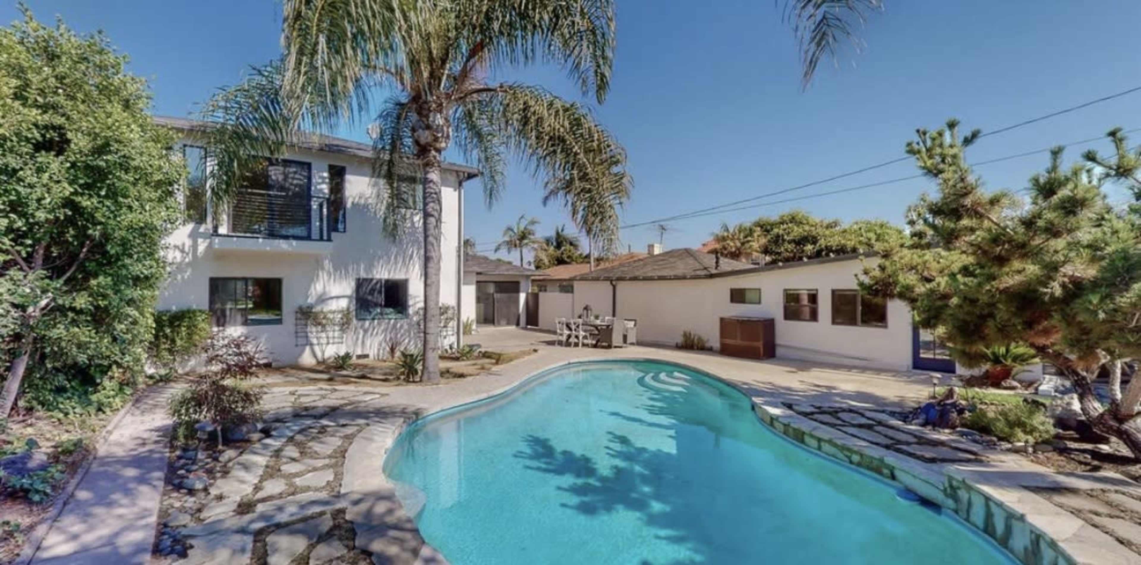 The image shows a backyard with a swimming pool surrounded by palm trees and a two-story house in the background.