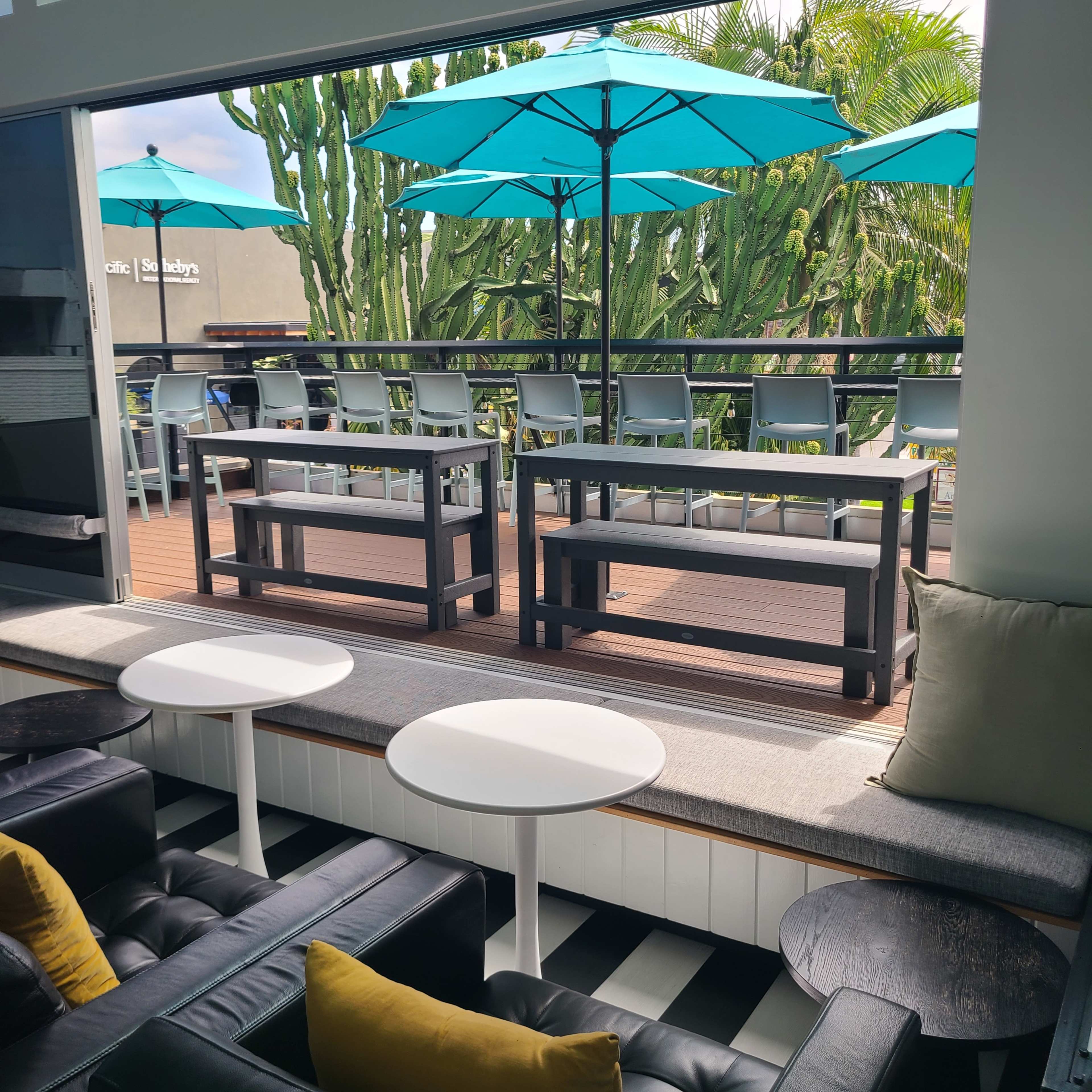 The image shows a modern seating area with black leather couches, small white tables, and a view of a patio featuring turquoise umbrellas and wooden furniture against a backdrop of cacti.