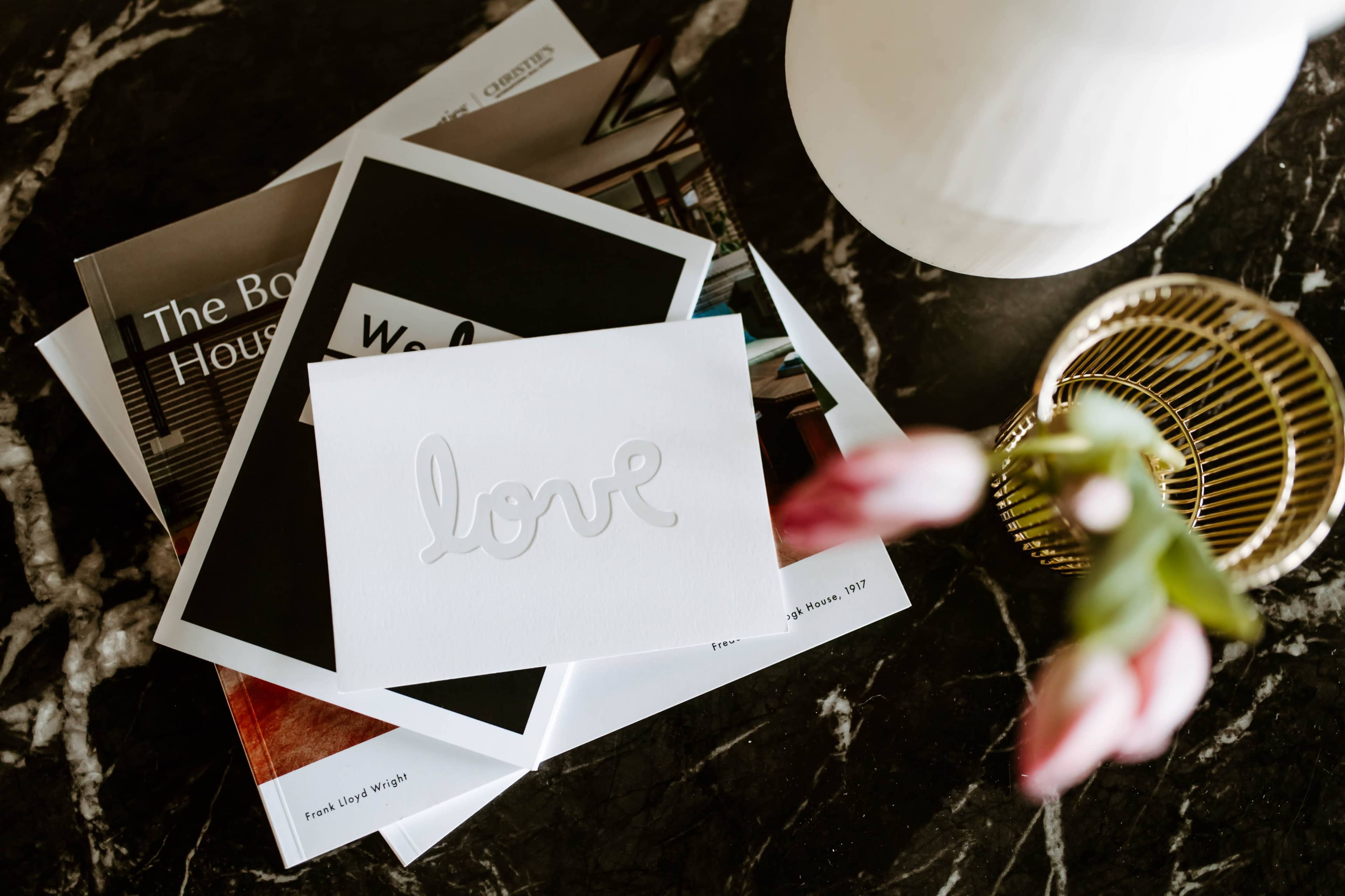 A white card with the word "love" embossed on it is placed on top of several magazines and a ceramic vase, alongside fresh flowers.
