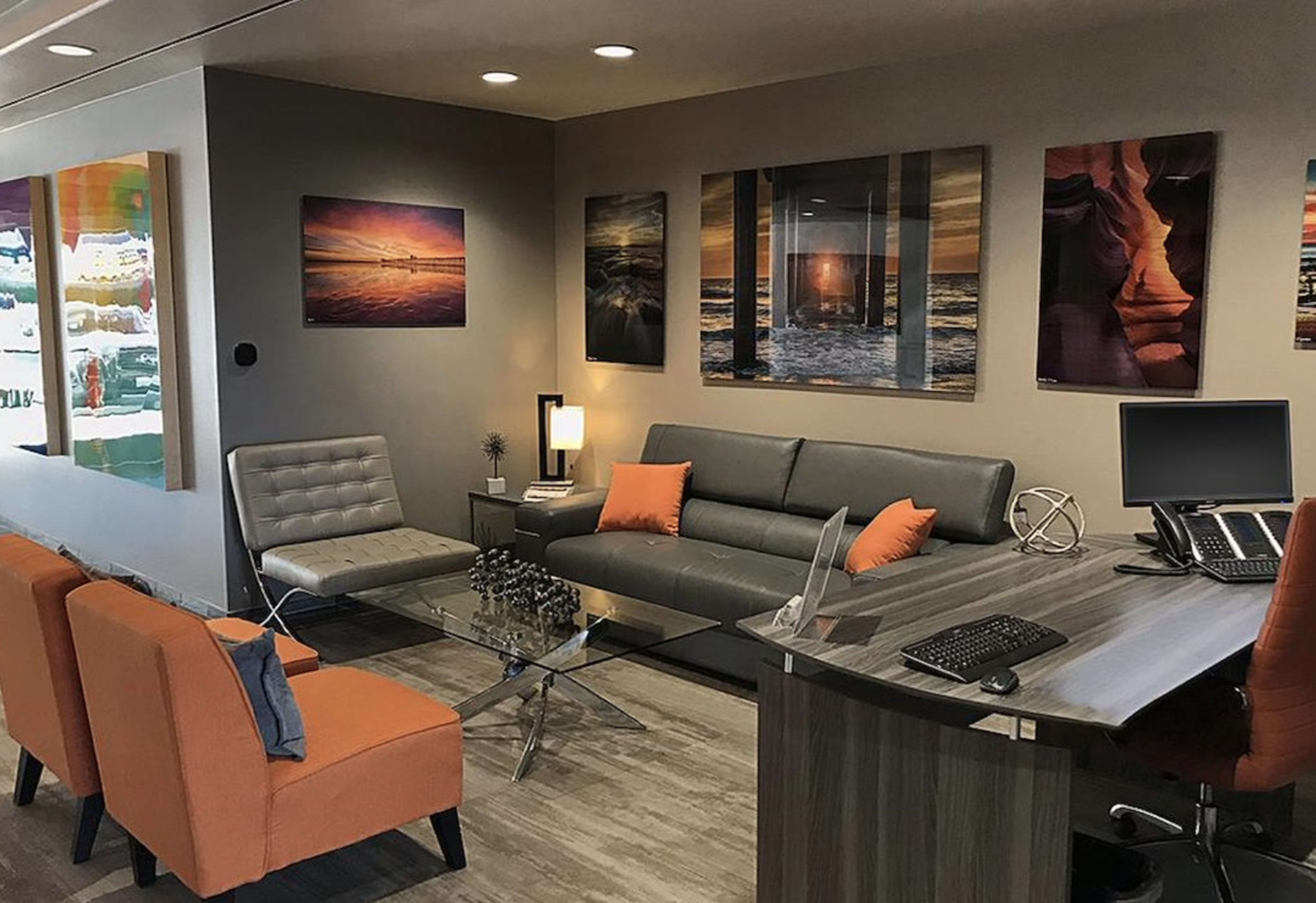 A modern waiting area featuring gray sofas, orange accent chairs, a glass coffee table, and artistic wall decorations.