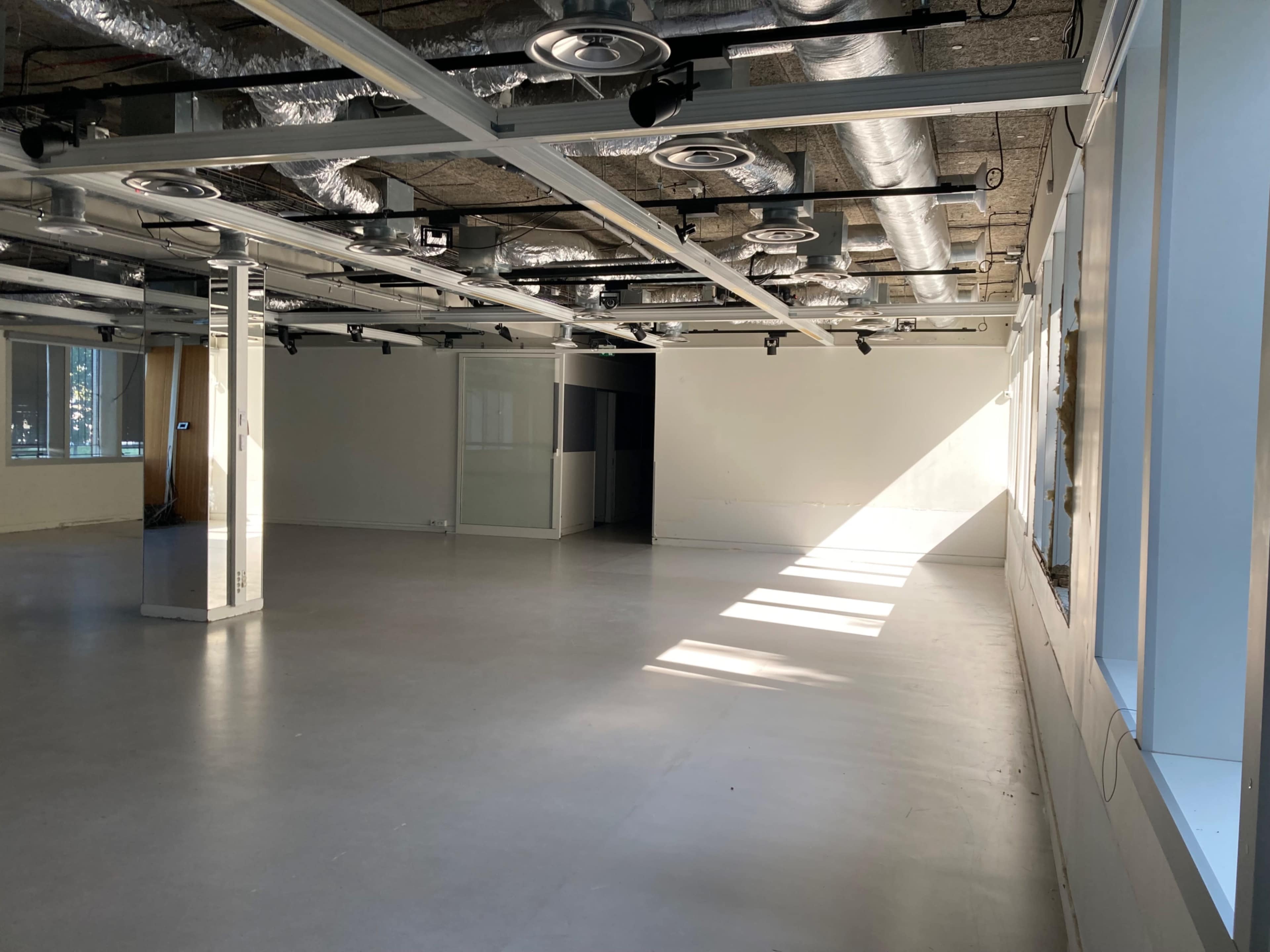 The image shows an empty office space with polished concrete floors, large windows allowing natural light, and exposed ductwork overhead.