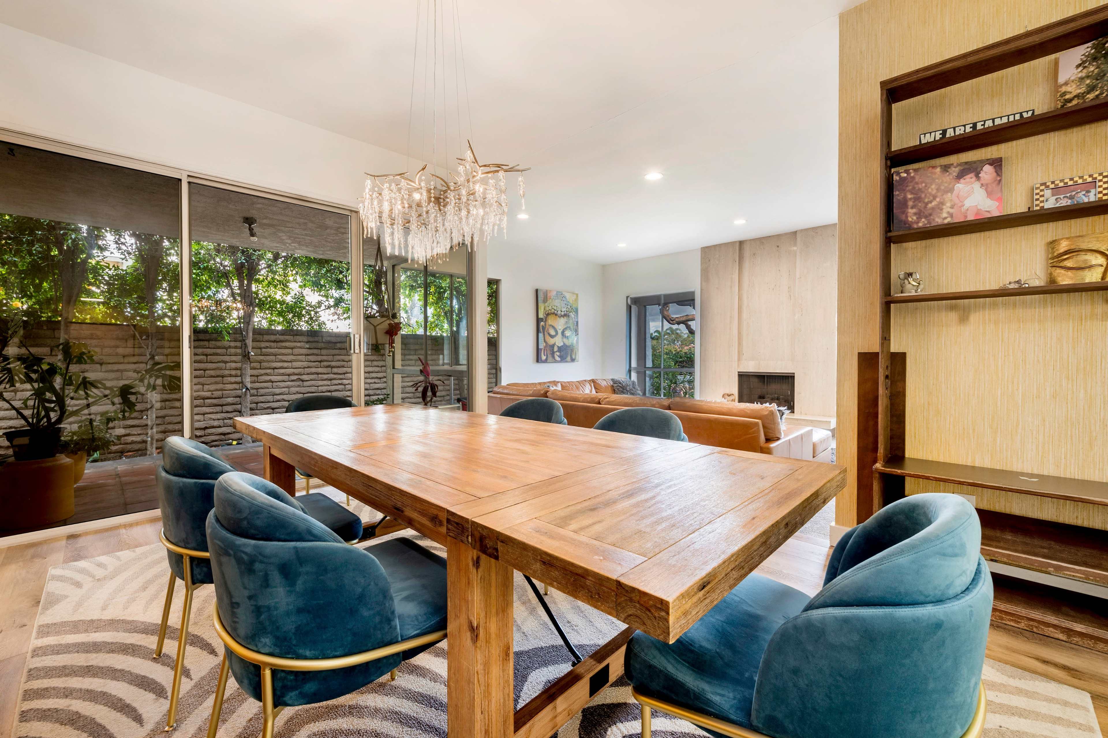 A modern dining area features a wooden table with blue chairs and a chandelier, surrounded by large windows and a view of a patio.