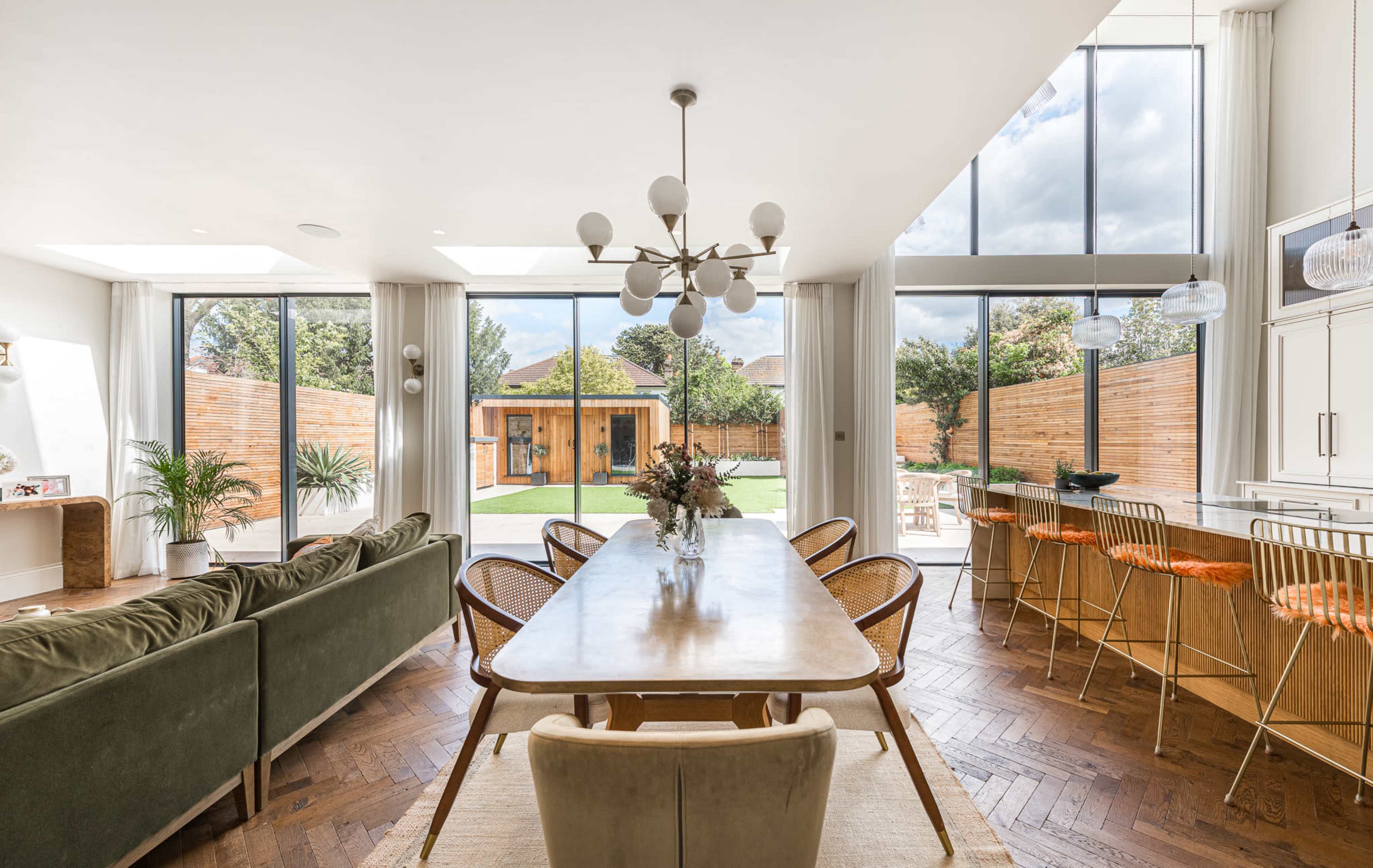 The image shows a modern, open-concept living and dining area featuring a long dining table, a plush sofa, large windows with natural light, and a view of a landscaped outdoor space.