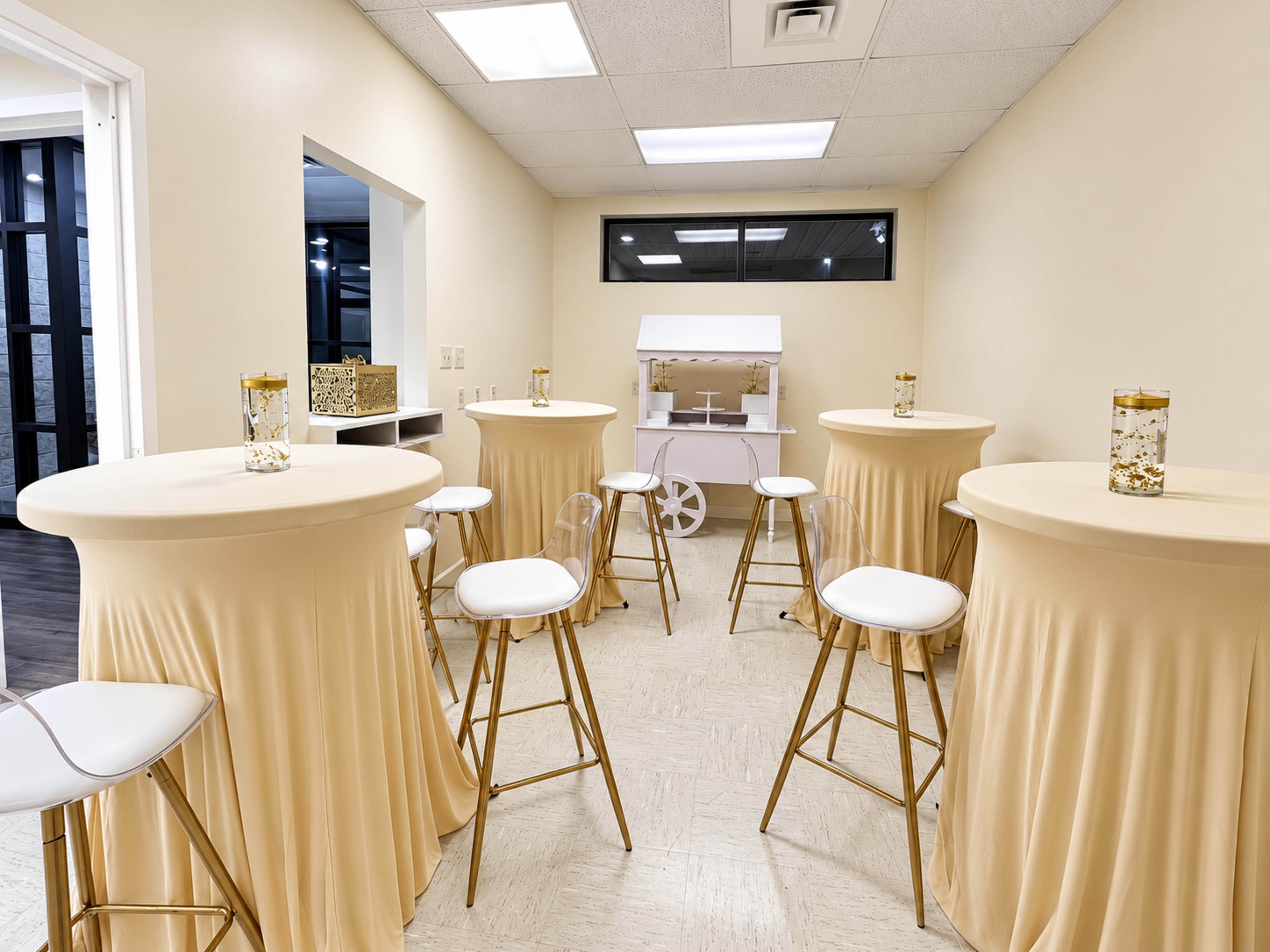 The scene features a bright, neutral-colored room with several tall round tables dressed in beige linens and surrounded by clear plastic chairs.
