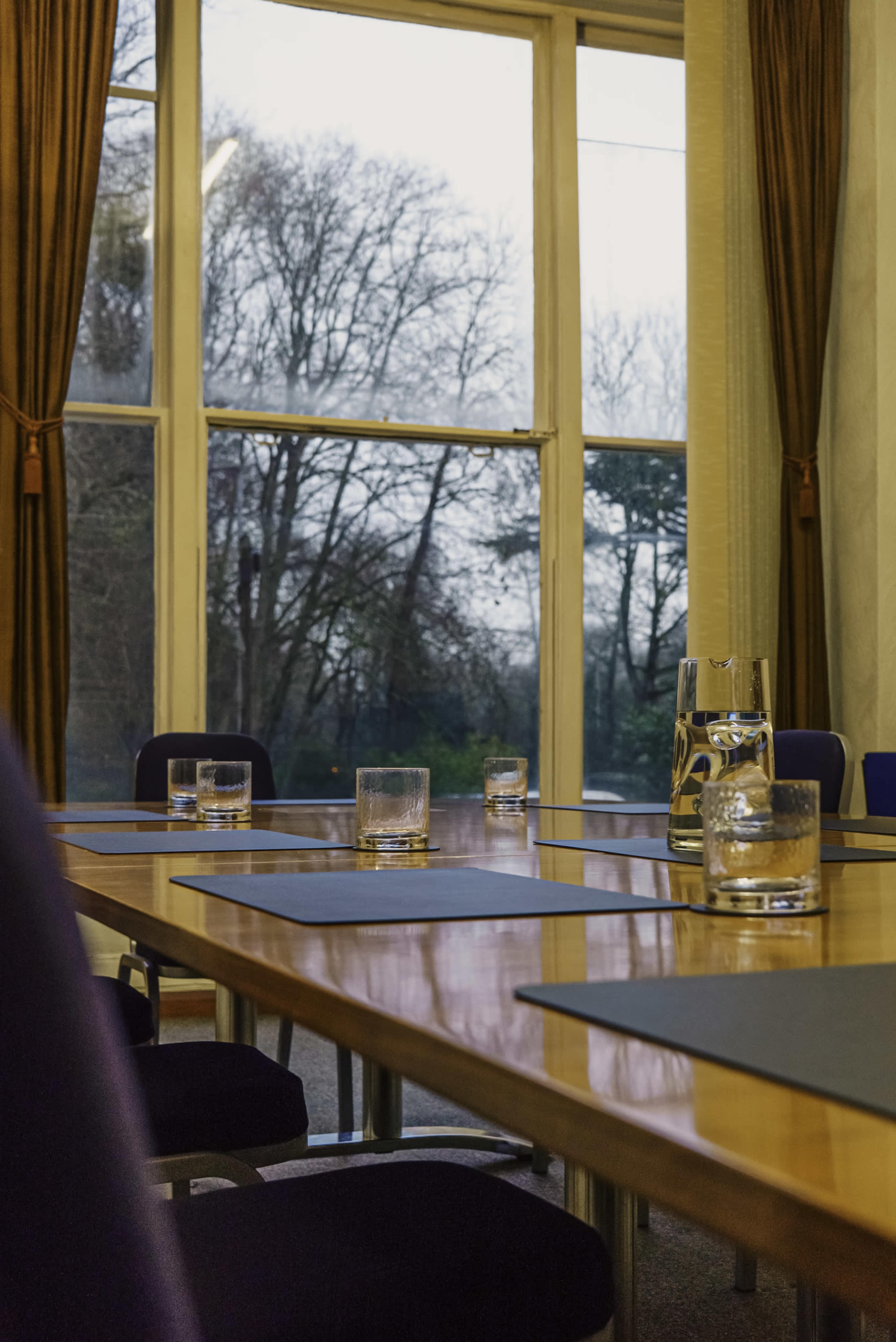 A conference room table with water glasses arranged on blue placemats overlooks a view of trees through large windows.