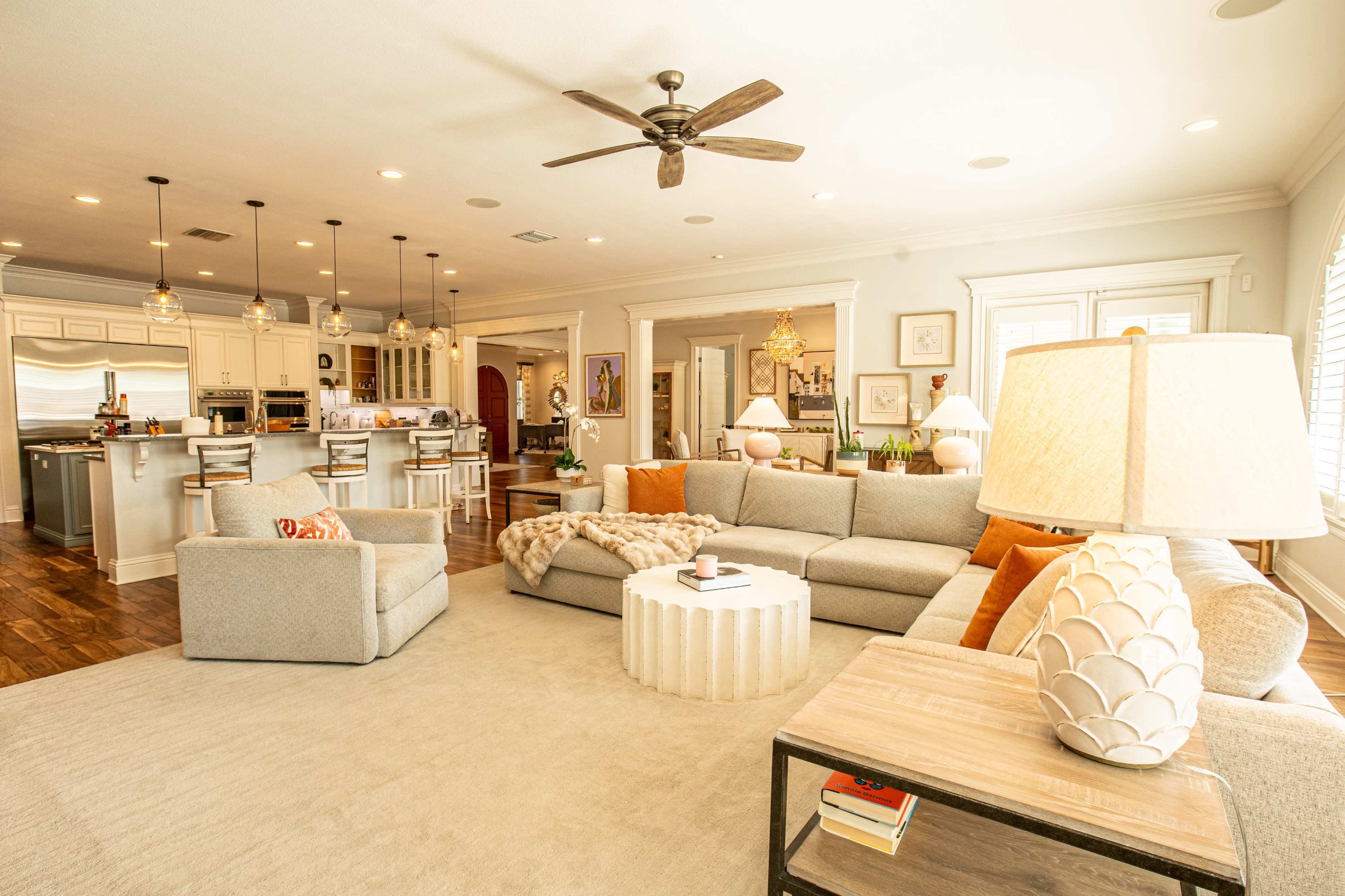 The image shows a spacious living room with a large sectional sofa, a round coffee table, a cozy area rug, and a view into an adjacent kitchen with a bar and dining area.