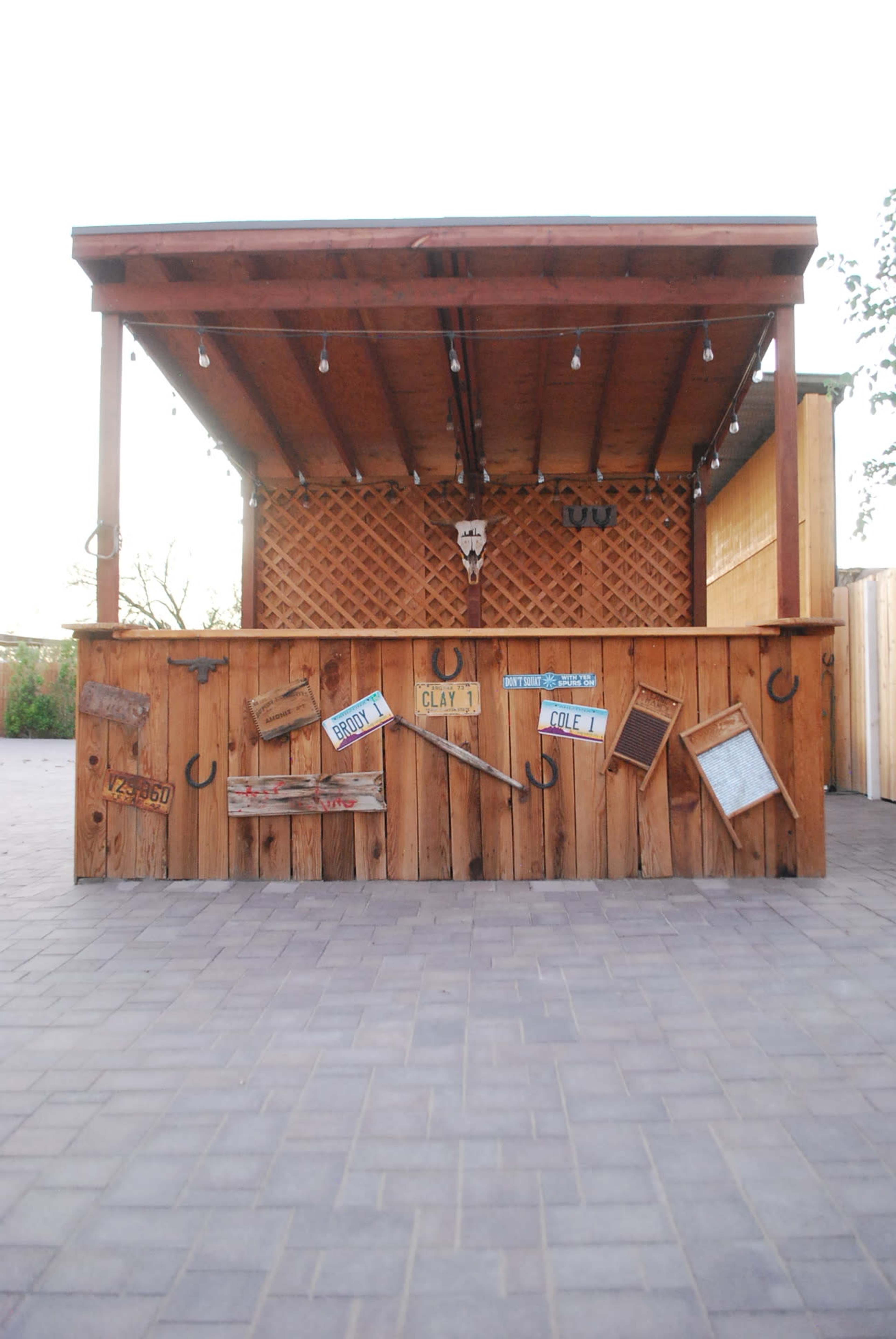The image shows a rustic wooden bar structure with a slanted roof, decorated with various items and signs, set against a clear sky.